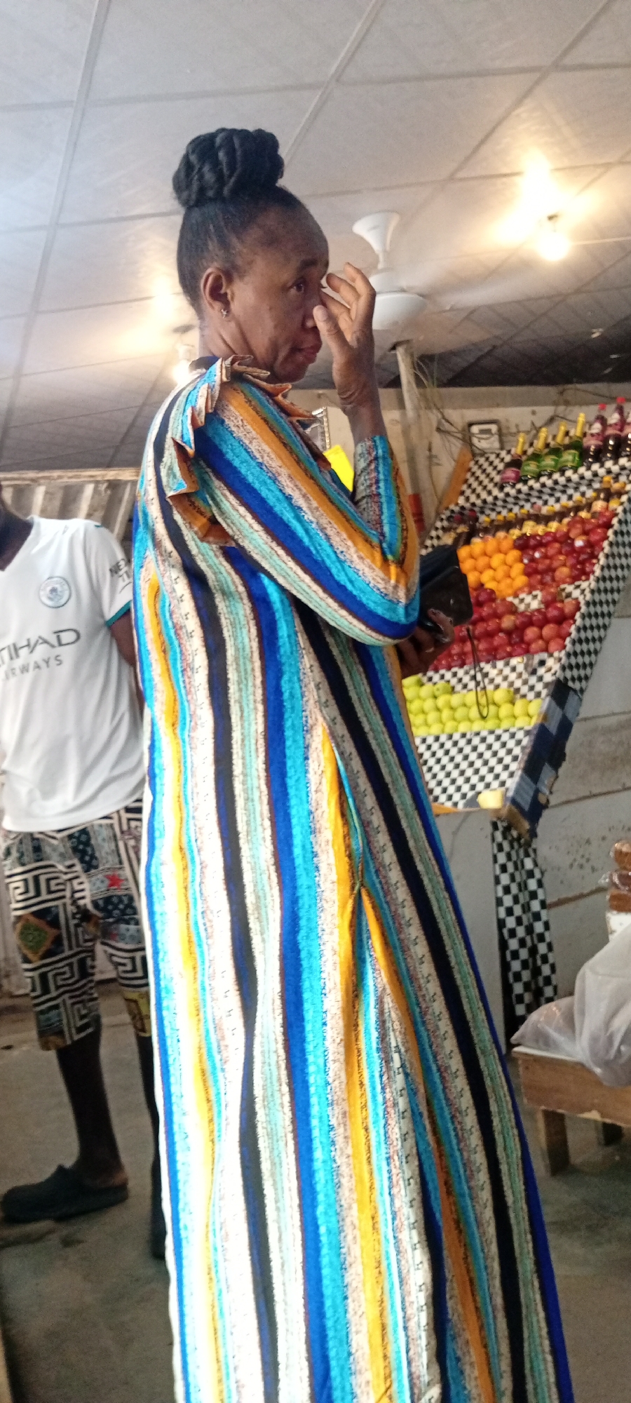 The image shows a woman standing in what appears to be a market or an outdoor shop in Aviation Quarters, Nigeria. She is wearing a long, brightly colored striped dress with blue, yellow, orange, and white stripes. Her hair is styled in a bun on top of her head. She is touching her face with her right hand, her expression is difficult to discern.
To her left, a person wearing a white t-shirt with "ETHAD AIRWAYS" written on it and patterned shorts is partially visible. In the background, a display of fruits, including oranges and apples, is arranged on shelves covered with a black and white checkered cloth. Some bottles, possibly beverages, are also visible on the top shelf. The scene appears to be outdoors, possibly during the daytime, with artificial lighting visible on the ceiling. The overall atmosphere seems to be that of a bustling marketplace.