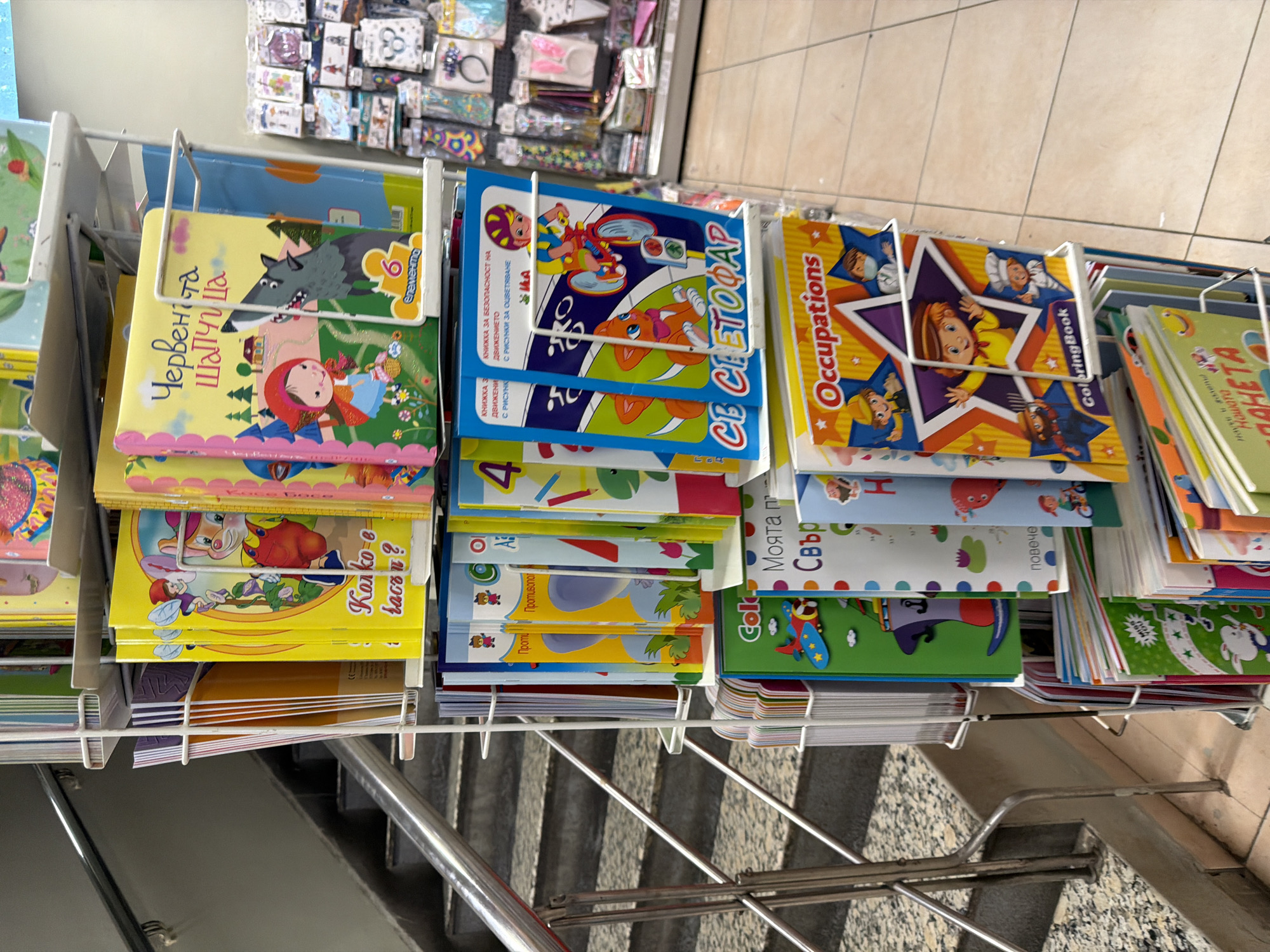 The image shows a retail display of children's books and activity books in a store. The books are organized on metal shelves. Some of the book covers visible include "Little Red Riding Hood" (Червената шапчица), a book titled "Coloring Book Occupations," and several books with colorful cartoon characters and Bulgarian text. There are also some smaller items, possibly accessories, displayed on shelves in the background. The environment appears to be an indoor retail space with tiled flooring. The location is identified as Pazardzhik, Bulgaria. No people are visible in the image.