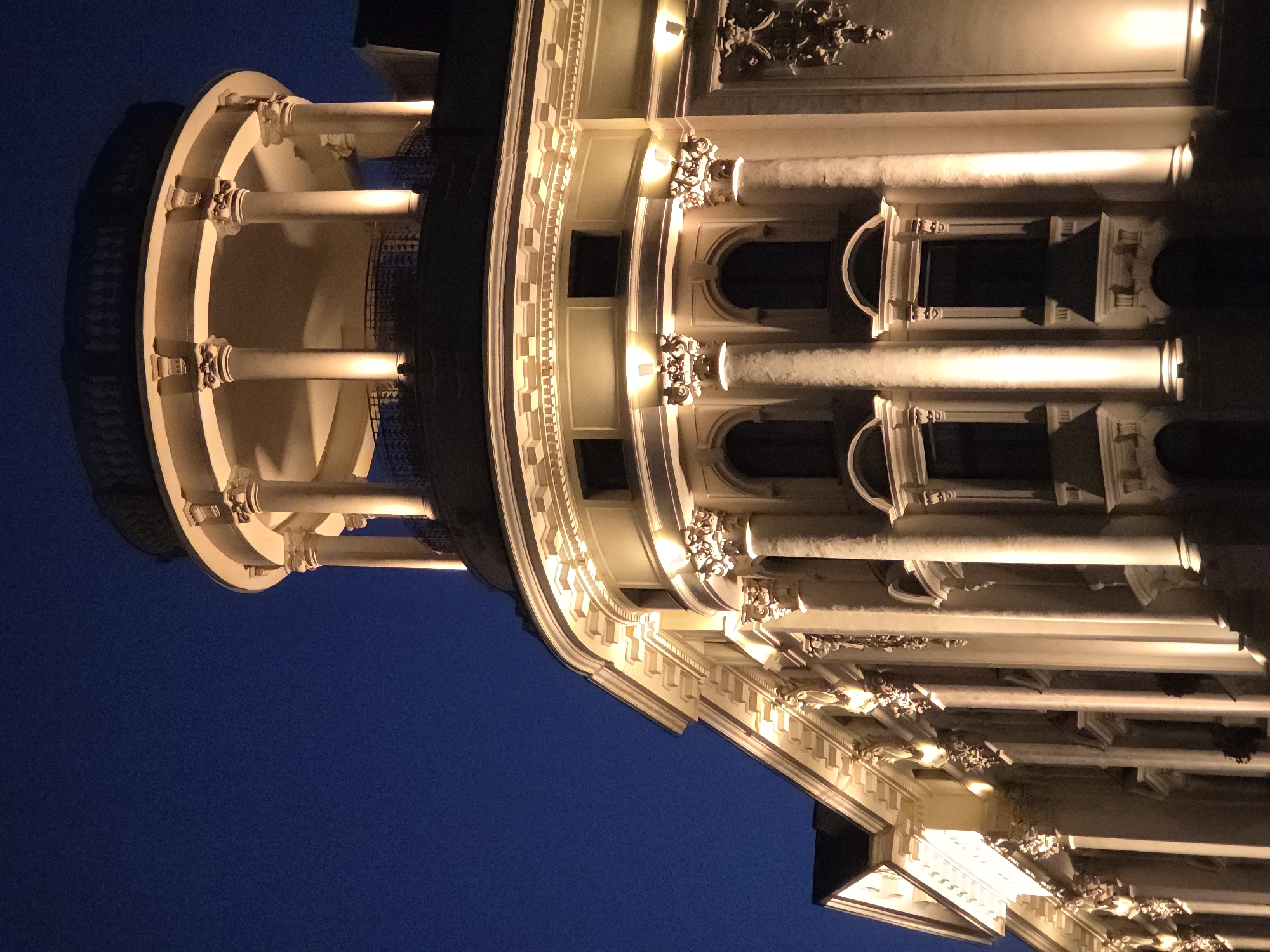 The image shows a portion of the building facades of the Presidential Palace in Warsaw, Poland, as seen from below at night. The architecture features ornate decorative elements including columns and intricate moldings illuminated by artificial light. A circular, colonnaded turret is on the left, while rows of windows and columns extend across the right side of the frame. The background is dark blue, suggesting the evening sky.