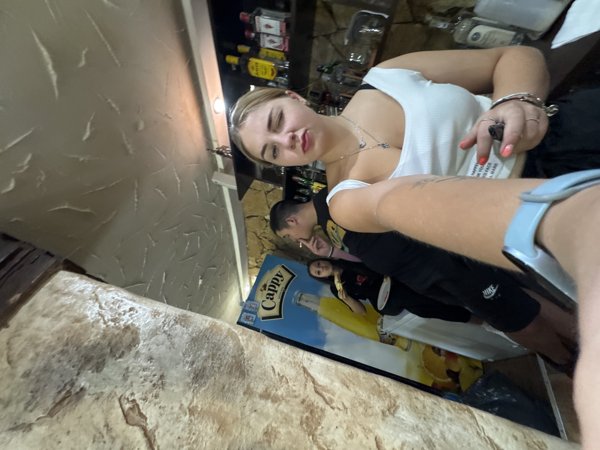 A young Caucasian woman with blonde hair, wearing a white tank top and black pants, is taking a selfie in what appears to be a bar or restaurant. She is winking and making a kissing face. Behind her, a man with short dark hair is smoking a cigarette and looking to the side. Another woman with dark hair is partially visible in the background, holding a slice of pizza. A large advertisement for "Cappy" juice is on display. Bottles of alcohol are visible on shelves in the background. The environment has textured walls and a textured ceiling. The specific location in the city of Sredets, Bulgaria, is not identifiable from the image.