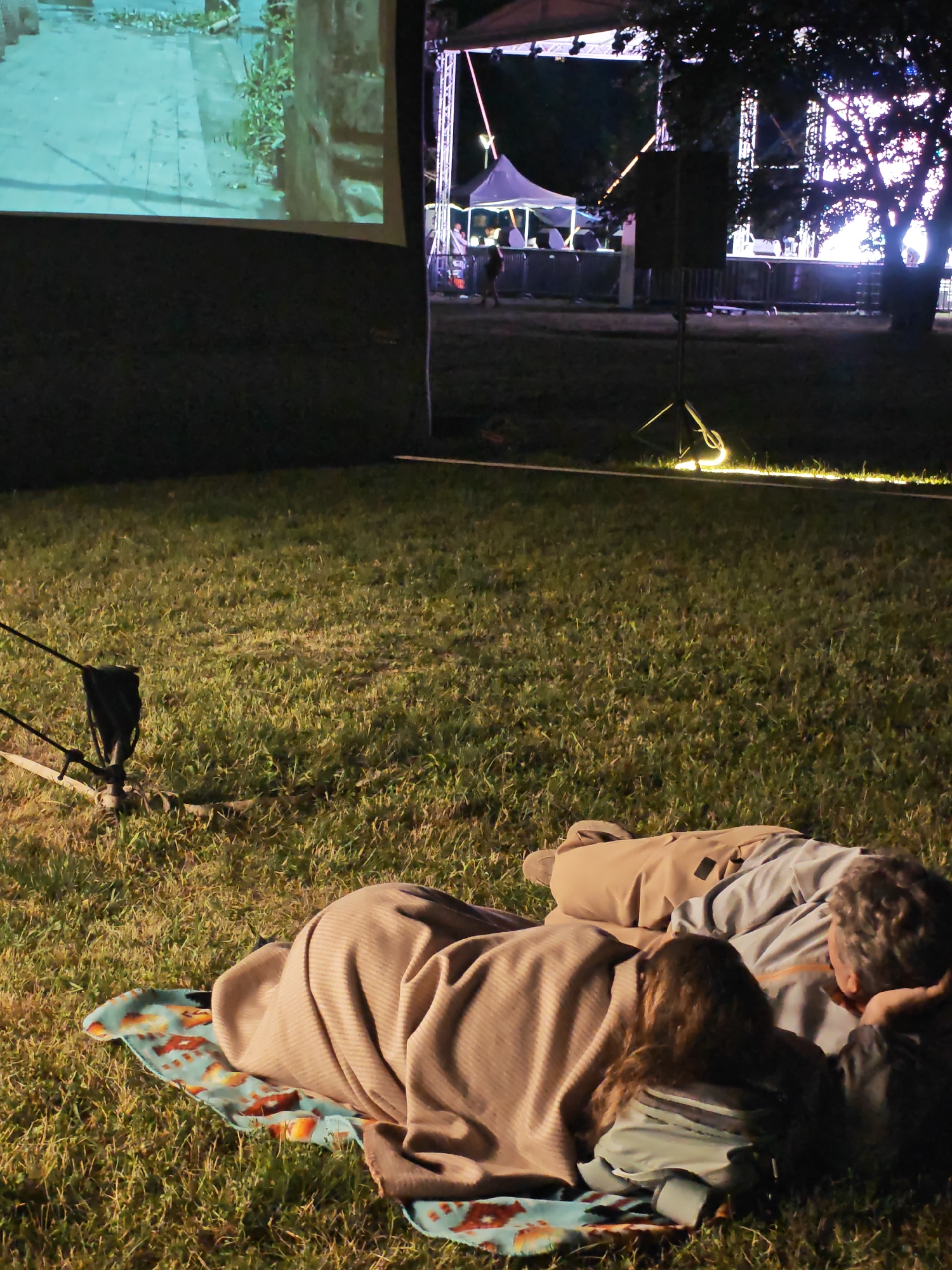 In an outdoor setting, two individuals are lying on the grass, covered by blankets. A projector screen is visible in the background, displaying an image of what appears to be a street scene with greenery. Beyond the screen, a stage is set up with lighting and structures, suggesting a performance or event is taking place. Several tents and barriers are also present in the background. The scene appears to be at dusk or night, with artificial lighting illuminating the area.