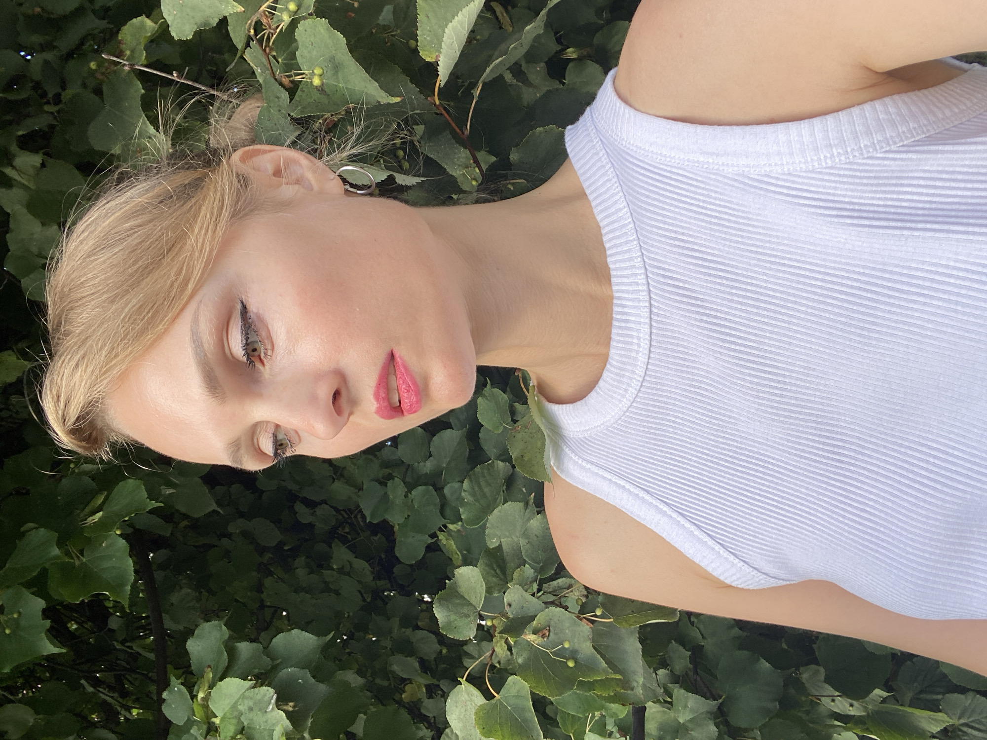 A blonde woman with bright pink lipstick and dark eyeliner is positioned in the foreground of the image, facing to the left. She is wearing a white ribbed tank top. Behind her is a dense backdrop of green leaves, suggesting an outdoor setting, likely within the city of Lida, Belarus, given the contextual prompt. The lighting appears natural, with sunlight filtering through the foliage.