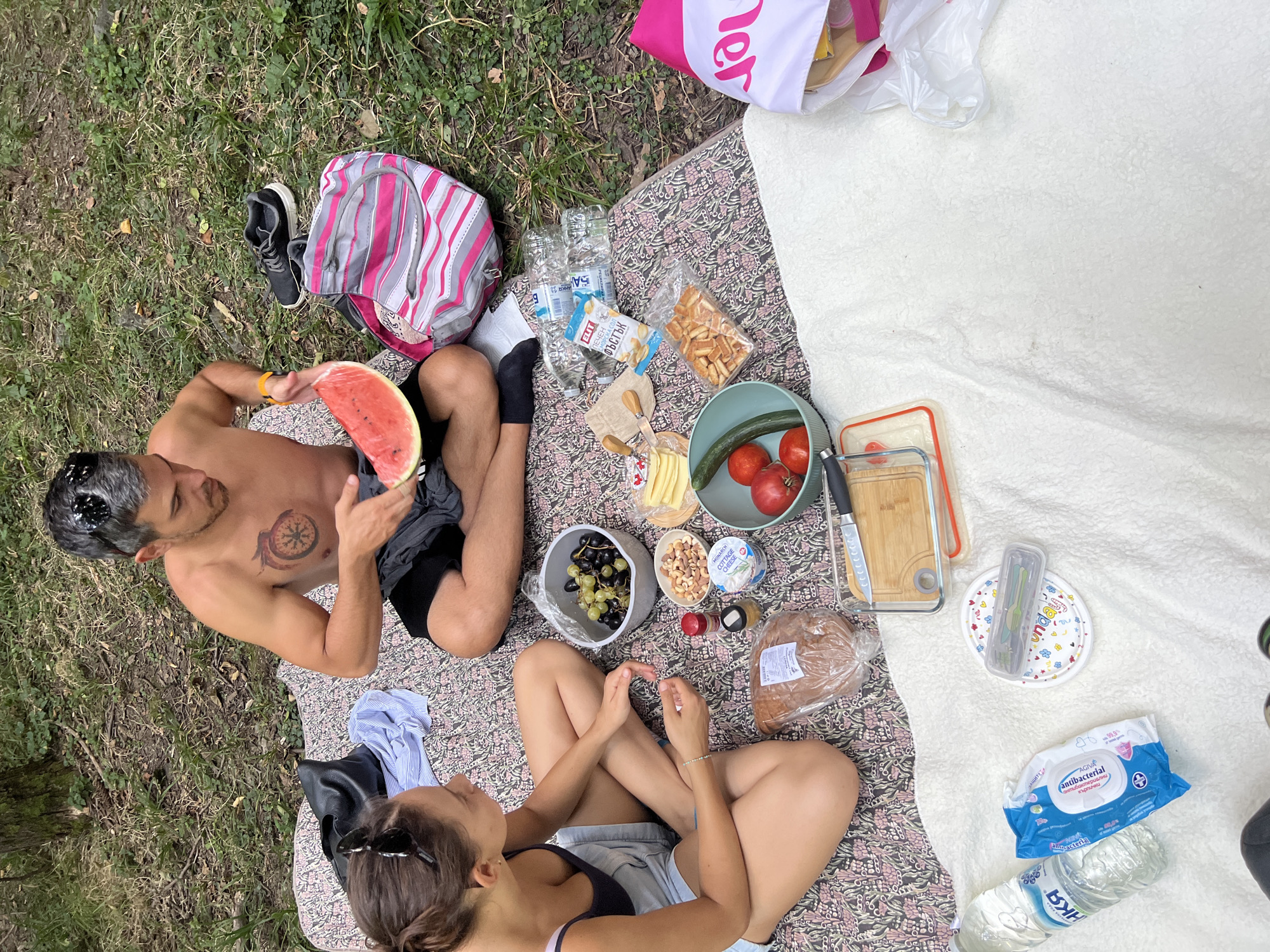 Two individuals are having a picnic on a blanket in a wooded area. The man, shirtless with a tattoo on his chest, is holding a slice of watermelon. A woman, wearing a dark top and denim shorts, is sitting opposite him, looking at the food. On the blanket, there is a variety of food items including grapes, nuts, cheese, bread, crackers, tomatoes, and cucumber. There are also water bottles, wet wipes, a picnic bag, and a pair of athletic shoes. The ground is covered with grass and fallen leaves, and a tree trunk is visible in the background. The scene suggests a casual outdoor meal in a natural setting, possibly a park or forest. The presence of trees and foliage indicates a rural or semi-rural environment. Without further context, it's impossible to definitively confirm the location as Zheleznitsa, Bulgaria, but the description aligns with a picnic setting that could exist in such a place.