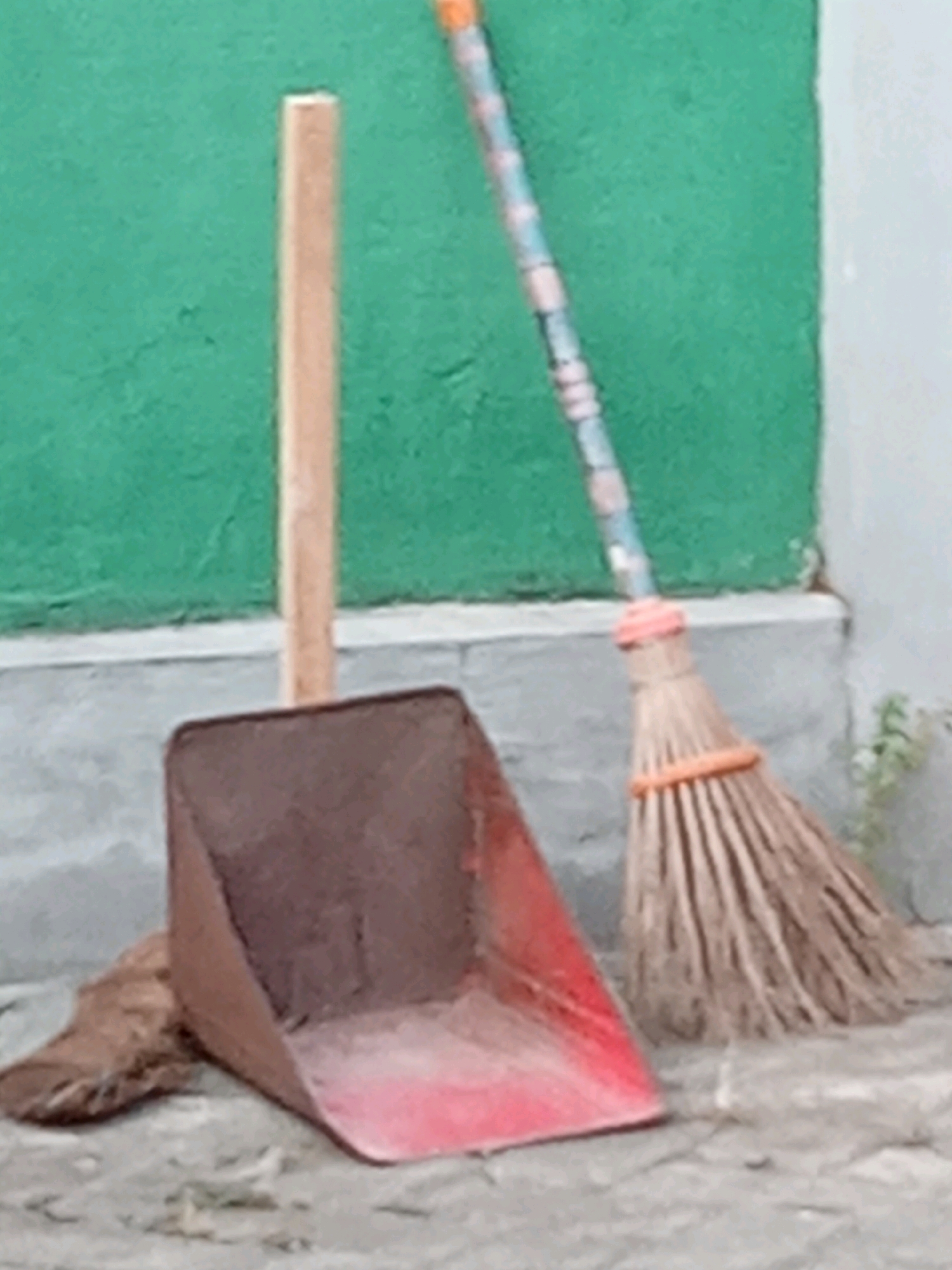 The image shows a red dustpan with a wooden handle leaning against a textured green wall and a gray concrete ledge. A broom with a colorful handle and natural bristles is leaning next to the dustpan, with its bristles resting on the ground. A brown, furry object, possibly a piece of cloth or an animal's tail, is partially visible in front of the dustpan. The ground is a cracked, gray surface. The scene is outdoors, likely in a simple outdoor area or alleyway in Sukoanyar, Indonesia, given the humble appearance of the cleaning tools and the wall. There are no people or visible actions taking place.