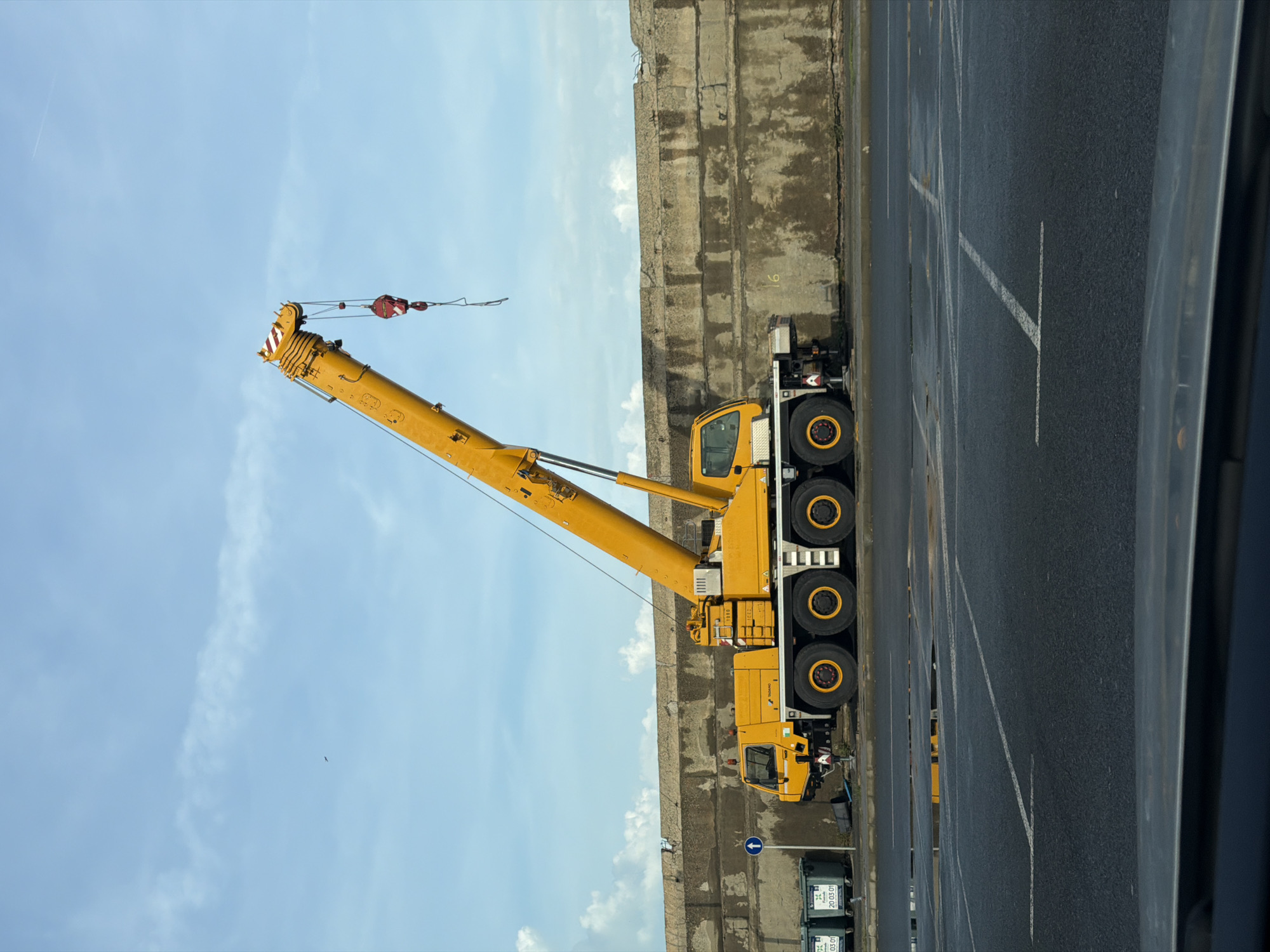 A yellow crane truck is parked next to a concrete barrier alongside a roadway in Burgas, Bulgaria. The crane boom extends diagonally towards a blue, lightly clouded sky. The truck is equipped with multiple axles and large tires. Two green garbage bins are visible near the truck's front. The roadway has white parking lines. The photo appears to be taken from an elevated position. The wall is gray stone.