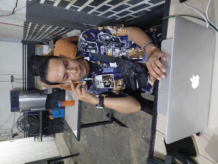 An indoor scene in Mns Alue, Indonesia, depicts an adult male seated at a light-colored table, facing the camera with a neutral expression. He is wearing a patterned short-sleeved shirt over a black t-shirt, dark trousers, a black fanny pack, a dark wristwatch on his left wrist, a silver-colored ring on his left ring finger, and multiple bracelets on his right wrist. His left index finger touches the skin below his left eye, while his right hand rests on the trackpad of an open silver Apple MacBook laptop in front of him. To the left of the laptop, a dark smartphone lies face down. To the right, another dark smartphone is connected by a white charging cable.
In the mid-ground, a second individual, wearing an orange or light brown t-shirt, is seated with their back to the camera at another dark-framed table with a light top. The background features a large dark industrial fan, a tall stainless steel machine with a digital display, and a light-colored display unit, possibly a freezer. An orange and blue plastic cup with a straw rests on the table behind the main subject. The walls are light-colored, with a horizontal black stripe running across the mid-section and a patterned area resembling black and white/grey bricks/tiles on the right. Exposed electrical wiring is visible on the walls. The floor is concrete.