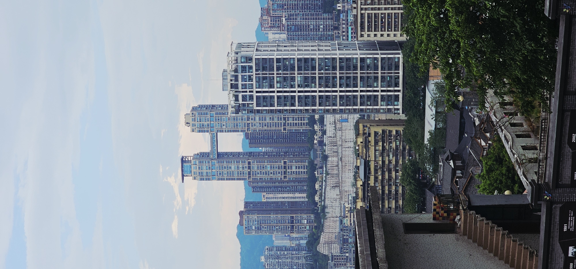 The image depicts a panoramic view of a dense urban landscape, likely in Yuzhong District, China, characterized by a mix of modern high-rise buildings and older, lower-rise structures. In the background, several prominent skyscrapers dominate the skyline, including a unique structure with two towers connected by a skybridge. The architecture suggests a contemporary city with significant development. In the foreground, a portion of a building with an exterior staircase and a textured facade is visible, leading the eye towards the dense city below. The middle ground features a cluster of mid-rise apartment buildings, appearing older than the skyscrapers, with visible balconies and window arrangements. Further down, traditional-looking roofs and trees suggest a contrasting architectural style or older neighborhood within the urban fabric. The overall environment is one of a bustling metropolis with varying architectural eras coexisting. The sky is partly cloudy, indicating daytime. There are no visible people or distinct actions taking place.