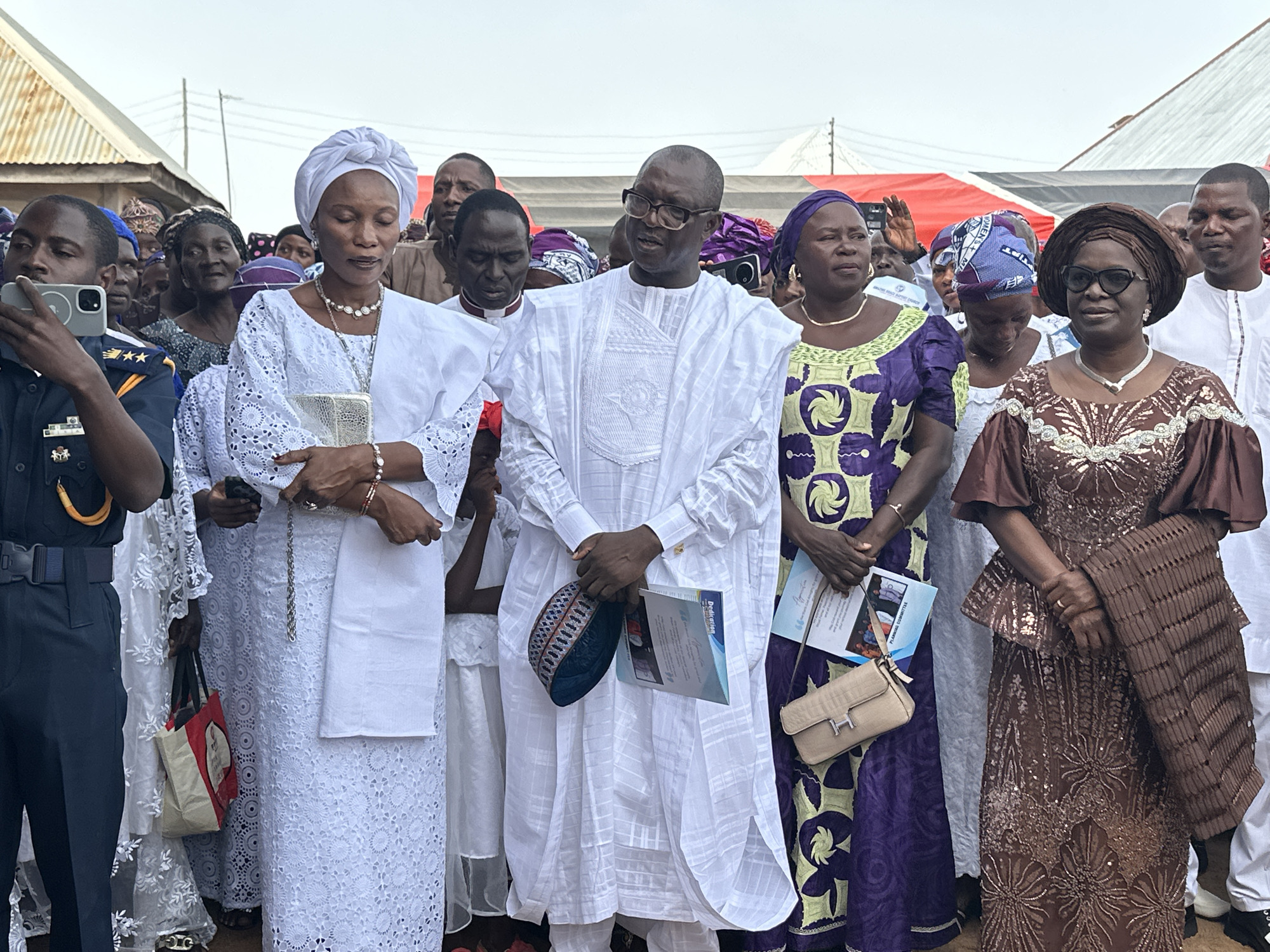 A group of people is standing together outdoors in Goni Gara, Nigeria. The central figure is a man wearing glasses and a white traditional garment, holding a dark blue patterned cap and a program titled "Dedication". To his left is a woman in a white lace dress and headwrap, holding a silver clutch. To his right, are two women in traditional dresses; one is wearing purple with yellow designs, and the other is wearing brown with sparkling accents. In the foreground, a man in a uniform is holding up a phone, seemingly taking a picture. Several other people, including some wearing traditional head coverings, are visible in the background. The setting appears to be in a village, with buildings visible in the background.