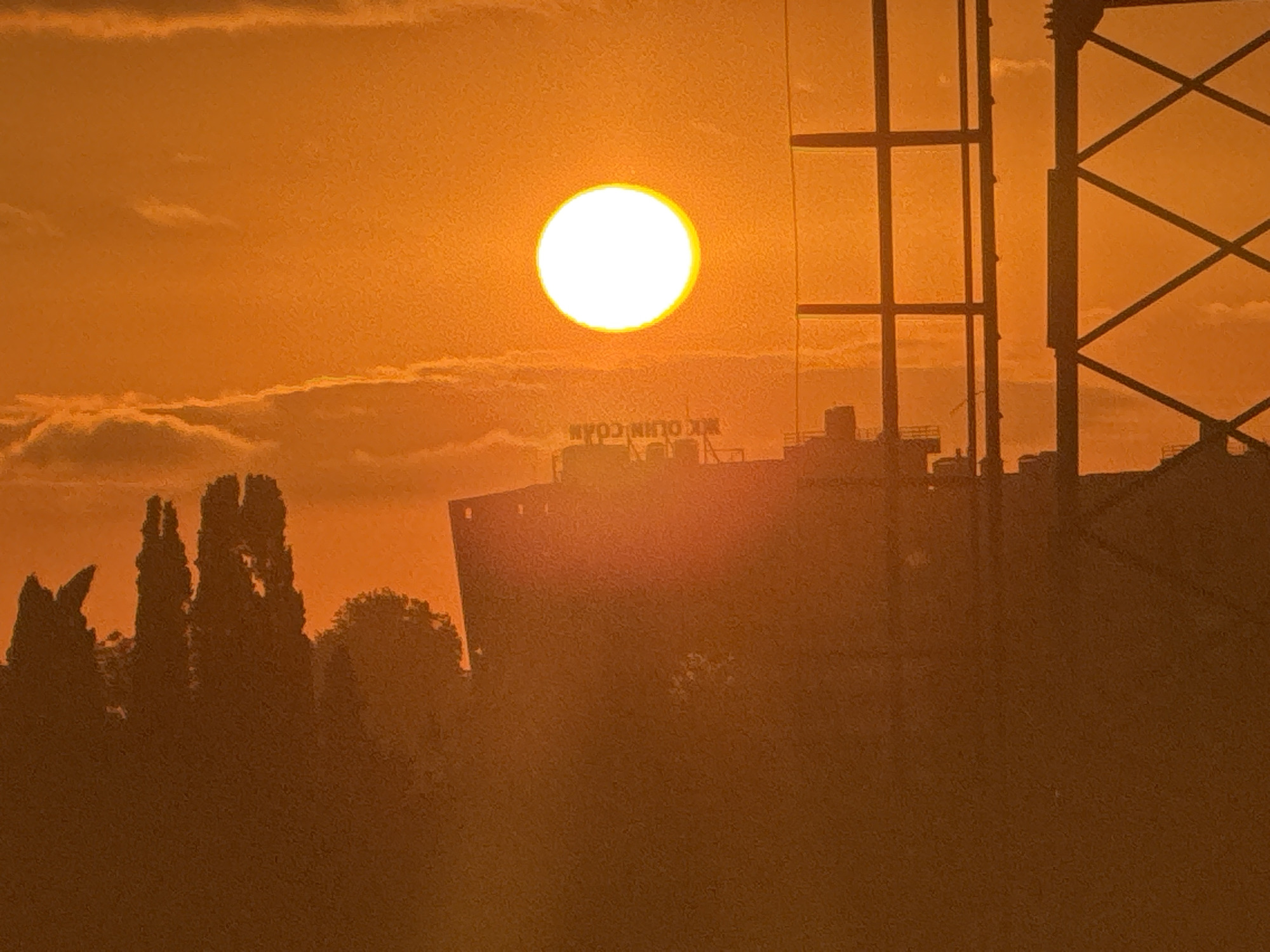 This media file depicts a sunset over Sochi, Russia. The sun, a bright white orb with a luminous halo, is positioned in the upper left quadrant of the frame, casting a warm, orange glow across the scene. Wispy clouds are visible, illuminated by the setting sun.
In the mid-ground, silhouetted against the sunset, is a large, flat-topped structure, likely a building or industrial facility. Above this structure, and slightly to the right, is a prominent, dark silhouette of a tall, lattice-like tower, possibly a radio tower or part of a construction crane. This tower creates a strong geometric contrast with the organic shapes of the trees in the foreground.
To the left of the main structure and extending towards the foreground are several dark silhouettes of tall, slender trees, characteristic of a cypress or similar evergreen species. The foreground is a dense, dark mass of foliage, suggesting a park or natural landscape.
The overall impression is one of a serene and possibly industrial landscape at dusk. The intense orange and yellow hues of the sunset evoke a sense of warmth and tranquility. The visible text, though partially obscured and reversed due to the angle, appears to read "MK OLIN COAH". The time of day is clearly sunset. The weather appears clear with some cloud cover.