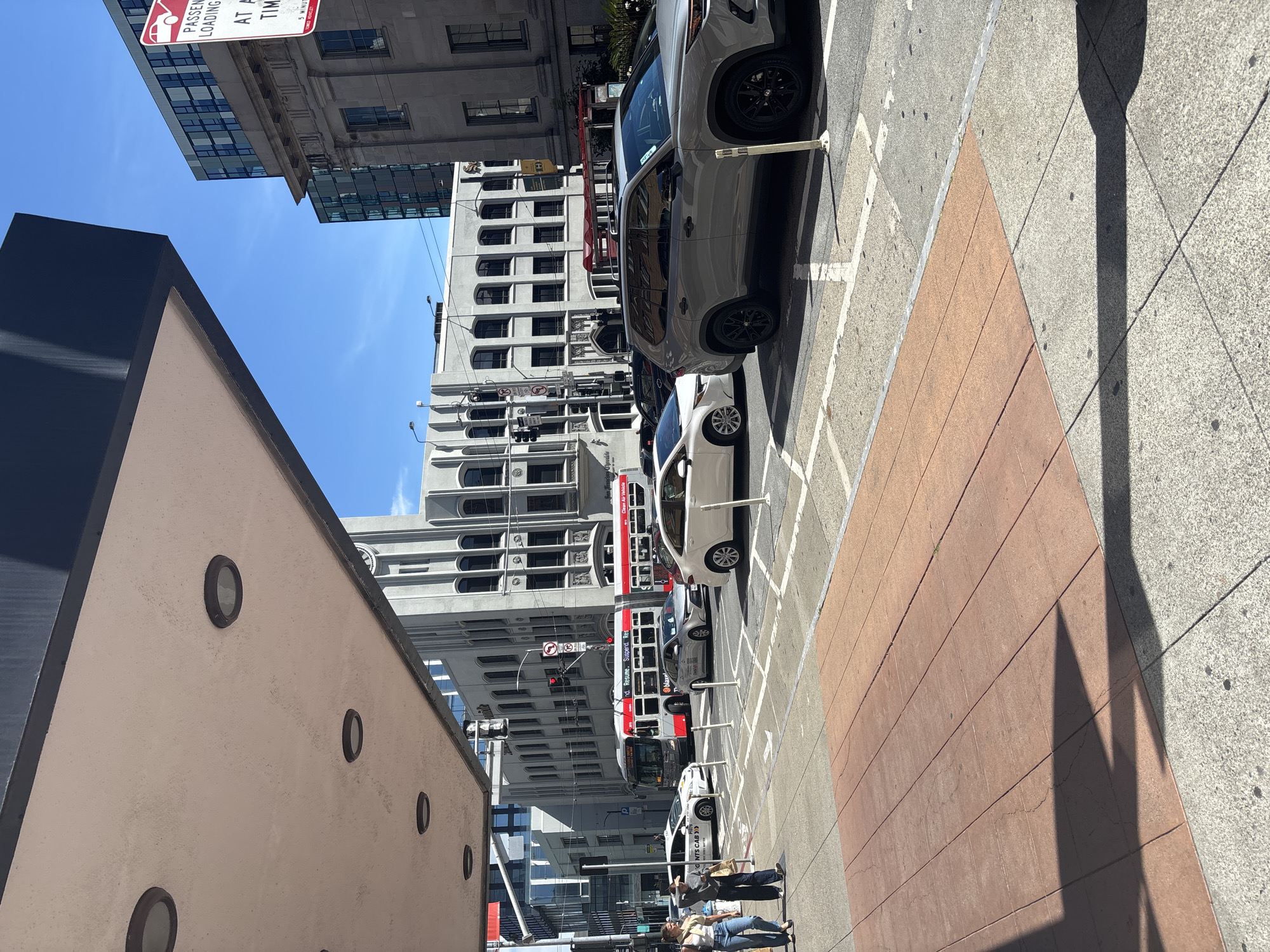 The image depicts a street scene in San Francisco, California, under a clear blue sky. In the foreground, a red brick sidewalk runs diagonally across the frame, transitioning to a gray, speckled sidewalk. A large overhang with circular lights extends from an unseen building on the left, casting a shadow on the sidewalk.
The street is bustling with traffic. A red and white bus with "20" displayed prominently on its front is stopped at what appears to be a bus stop. Several cars are visible on the road, including a white sedan, a silver sedan, and a gray SUV. Pedestrians are present on the sidewalk, two women walking together.
In the background, various buildings line the street. Some have an older, ornate architectural style with grey stone facades and arched windows, while others are modern glass and steel structures. A red and white sign for passenger loading is visible on the right side of the frame. The overall impression is of a vibrant urban environment in a major city.