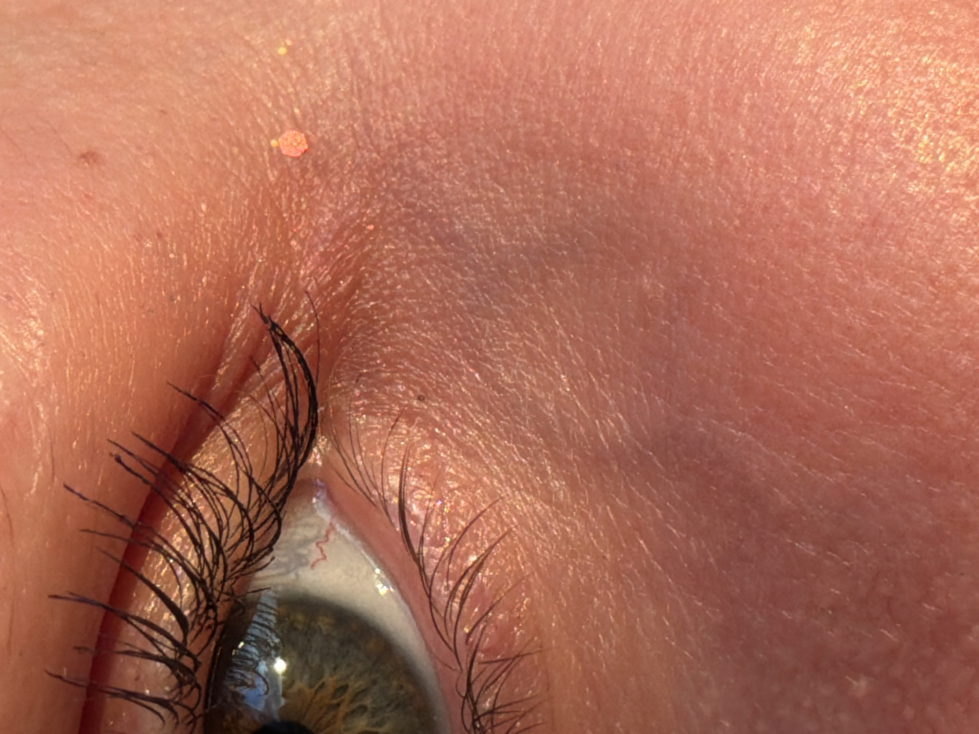 This is an extreme close-up, macro photograph of a human eye and the surrounding skin. The eye is open and looking to the left. The iris is a mix of green and brown with visible speckles. The eyelashes are long and dark, some appearing clumped together, possibly due to mascara. The skin is a warm, peachy tone and appears to have a subtle shimmer or glitter on its surface, particularly around the eyelid and cheek area. There are fine lines and pores visible in the skin texture. The lighting suggests it might be natural sunlight, as there are highlights and reflections on the skin and within the eye. No specific location in the city of Chaikovsky, Russia can be determined from the image.