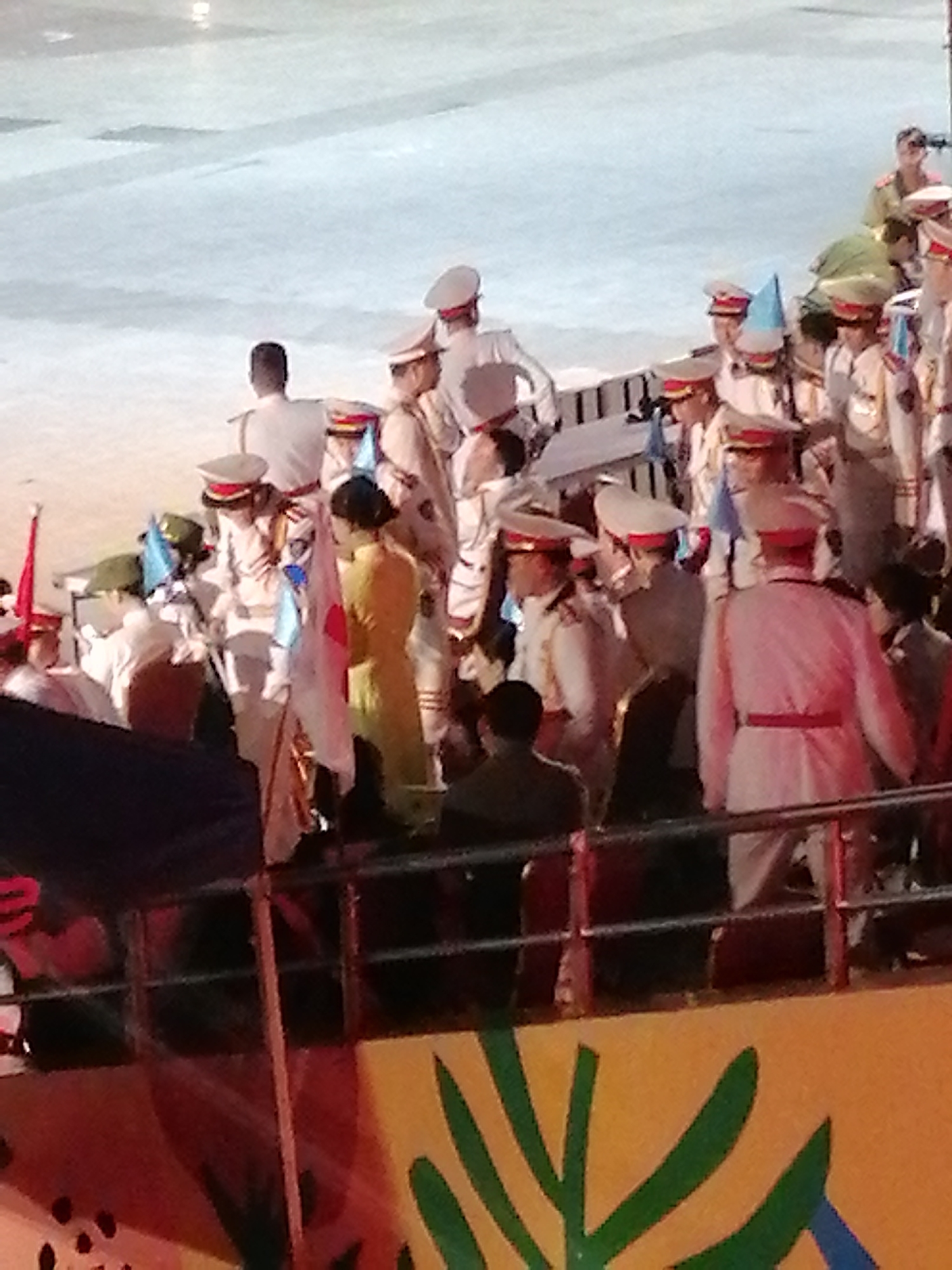 A group of people, many in military-style uniforms, are gathered on a light-colored surface that could be a stage or an outdoor event area. The uniforms are predominantly white with red trim and caps, while some individuals wear darker, olive-green uniforms. A few people are seen holding flags, including a white flag with a red circle and a Japanese flag. One person in the foreground is wearing a yellow outfit. The scene appears to be an outdoor event, possibly a parade or a formal ceremony, given the attire and the flags. The lighting suggests it could be daytime, though the specific time of day is unclear. The overall atmosphere seems formal and organized, with no overt signs of distress or celebration. The background is blurry, but hints at structures or a crowd. The foreground shows a railing and colorful decorations, with green leaf-like shapes and black dots on an orange background. There is no visible text in the image. The setting is identified as Da Lat, Vietnam.