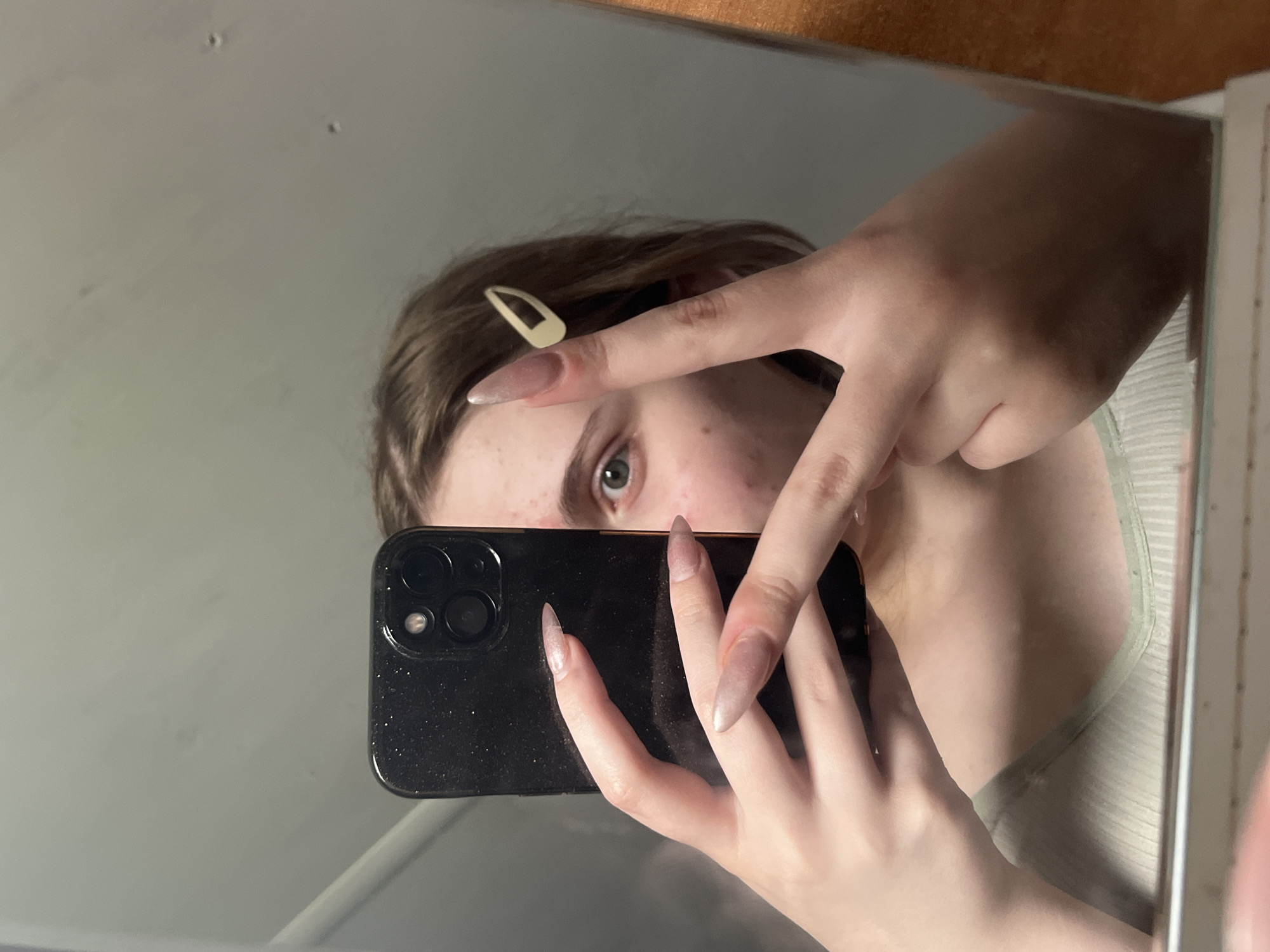 The image shows a young person holding a black smartphone in front of their face, taking a mirror selfie. They are wearing a light-colored, ribbed top. Their right hand is raised, with two fingers extended in a peace sign, and their left hand is holding the phone. They have long, light-colored fingernails. A light-colored hair clip is visible in their brown hair. The background appears to be a wall, with a mirror reflecting the scene. The lighting is dim. There is no visible indication of the city of Ashkadar, Russia, or any other specific urban or geographical location.