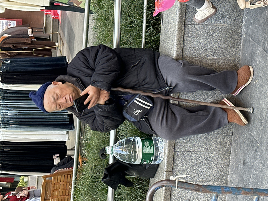 An East Asian male, appearing elderly, is seated on a grey stone ledge in Jiangjin, China. He wears a dark blue knit beanie, a black puffer jacket, dark grey sweatpants, and brown slip-on shoes with striped soles. He is holding a black smartphone with both hands, looking down at the screen with his eyes partially closed. A wooden walking stick rests between his legs, touching the paved ground. A black and white shoulder bag with a logo is visible across his torso. To his left, on the ledge, sits a large clear plastic water bottle with a green label and a black fabric bag. Behind the ledge is a green hedge. In the background, multiple clothing racks display various garments, suggesting an outdoor market setting. A person in a red top is partially visible on the far left, and a child's legs and feet, along with a pink object, are partially visible on the far right. A blue metal frame is present in the lower left foreground.