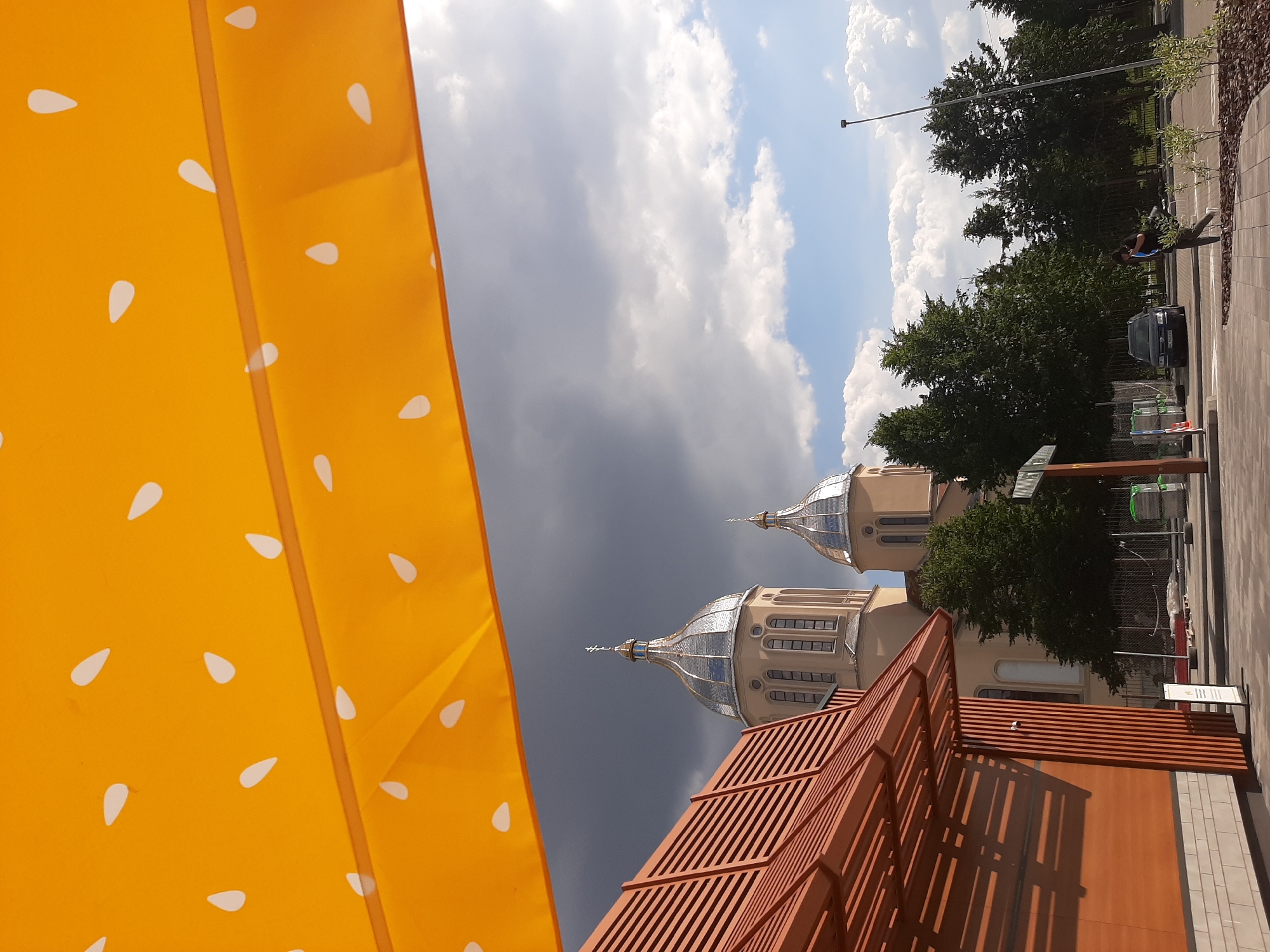The image captures a view of an architectural structure with domes against a dramatic sky. In the foreground, a bright orange surface with a white polka dot pattern, possibly a tent or awning, dominates the left side of the frame. To the right, two ornate domes with silver, reflective surfaces rise above a light-colored building. The sky is a mix of dark, stormy clouds and patches of bright blue with white cumulus clouds. Trees and a street with pavement are visible behind the building.
In the background, a person in dark clothing is seen near a parked dark car, walking along the street. There are fences and some construction or landscaping elements. A lamppost is also visible. The overall impression is one of an urban setting with an interesting juxtaposition of natural elements (sky, trees) and man-made structures. The time of day appears to be daytime, with the weather indicating a potential for rain due to the dark clouds. The location is identified as Lviv, Ukraine. No discernible text is visible in the image.