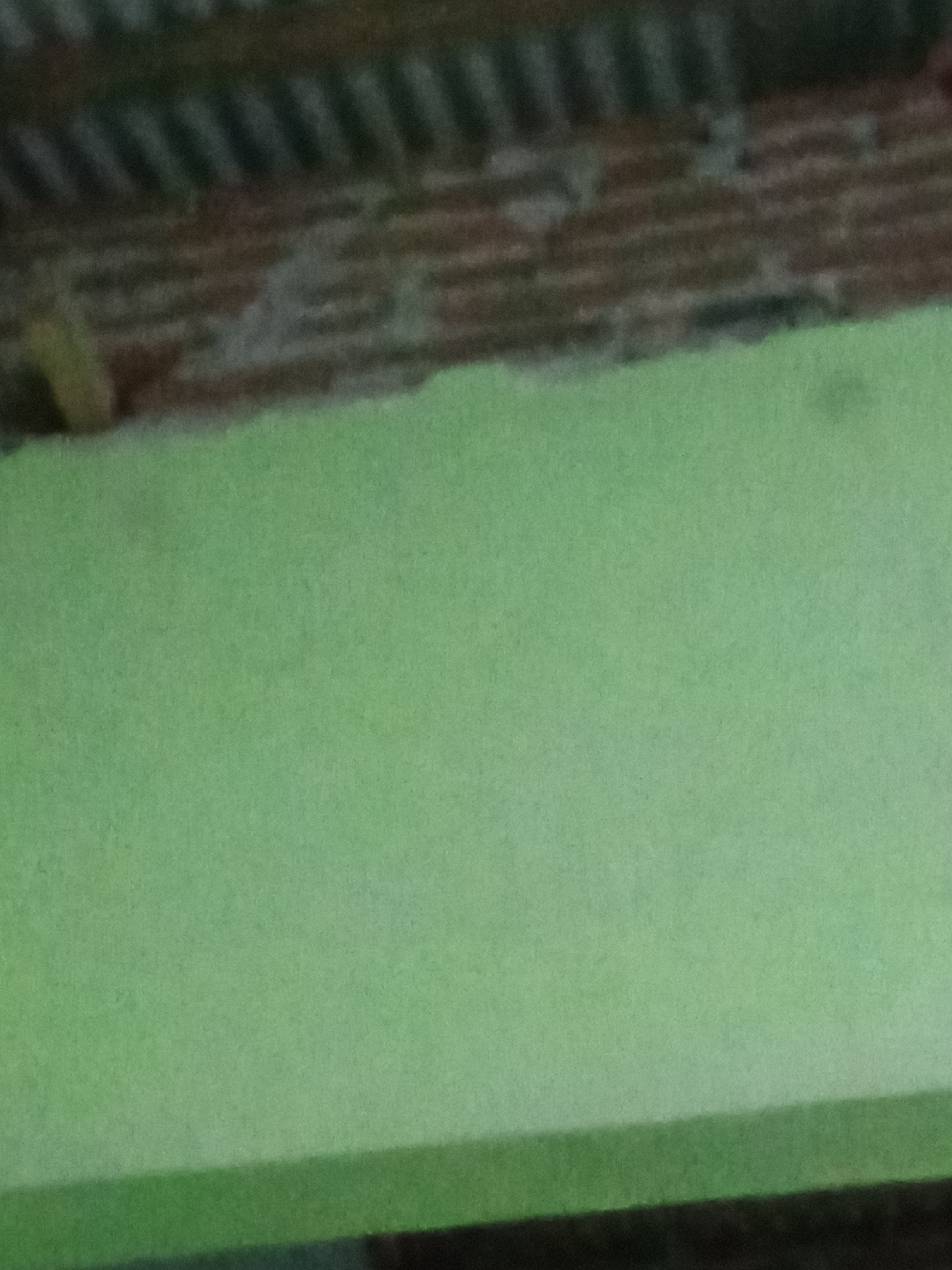 The image displays a blurry, close-up view of a wall and a roof structure. The wall is painted a light green color. Above the green wall, there is a section that appears to be a roof, made of dark, possibly weathered material with visible horizontal lines or corrugations. A small portion of what seems to be a yellow or light brown object is visible at the top left edge of the image, possibly protruding from the roof structure. The lighting is diffuse, and there are no discernible people or specific activities taking place. The overall impression is that of an outdoor, possibly construction or weathered facade, environment. The image does not provide any specific visual cues to pinpoint a location in Pemalang, Indonesia.