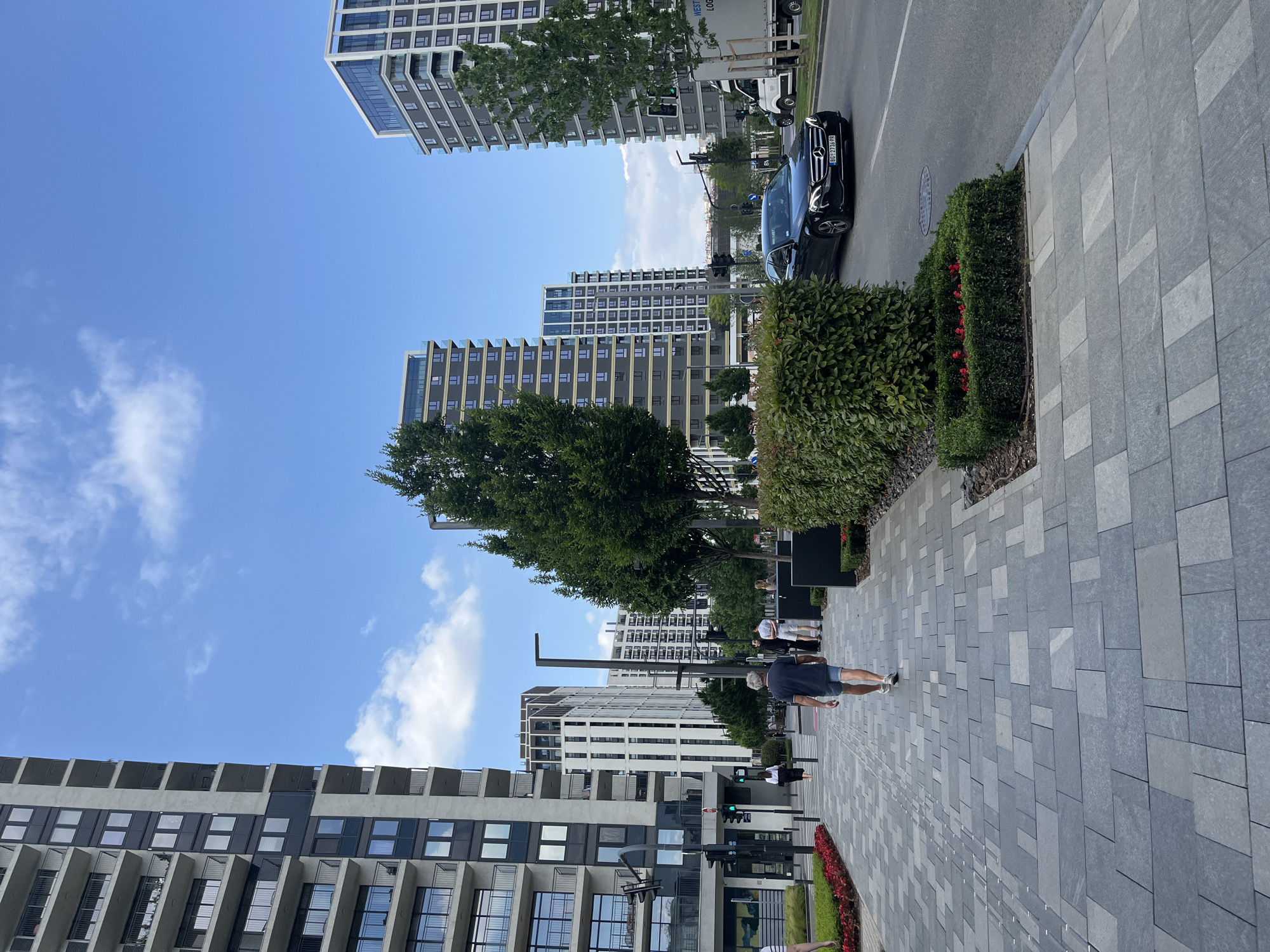 The image displays a modern urban street scene in Belgrade, Serbia, under a clear blue sky with a few wispy white clouds. The setting appears to be a newly developed area, characterized by contemporary high-rise buildings with distinctive architectural styles, featuring large glass windows, balconies, and a mix of light and dark grey facades.
A wide pedestrian sidewalk, paved with a pattern of grey and lighter-toned square stones, occupies the right side of the frame. Along the sidewalk's edge, well-maintained landscaping includes neatly trimmed green bushes and planters with vibrant red flowers. Several green trees are strategically placed, offering shade and natural elements amidst the urban structures.
On the asphalt road running parallel to the sidewalk, a black Mercedes-Benz sedan is visible, identifiable by its license plate "BG 2779PR," which indicates Belgrade. Further down the street, a white truck with "WEST LOG" printed on its side is partially seen.
Several people are present on the sidewalk. In the mid-ground, a man wearing a dark t-shirt and light shorts is walking away from the camera. Other pedestrians are scattered further along the path. The scene is bright, suggesting daytime, and the overall atmosphere is calm and orderly, reflecting a well-maintained urban environment.
