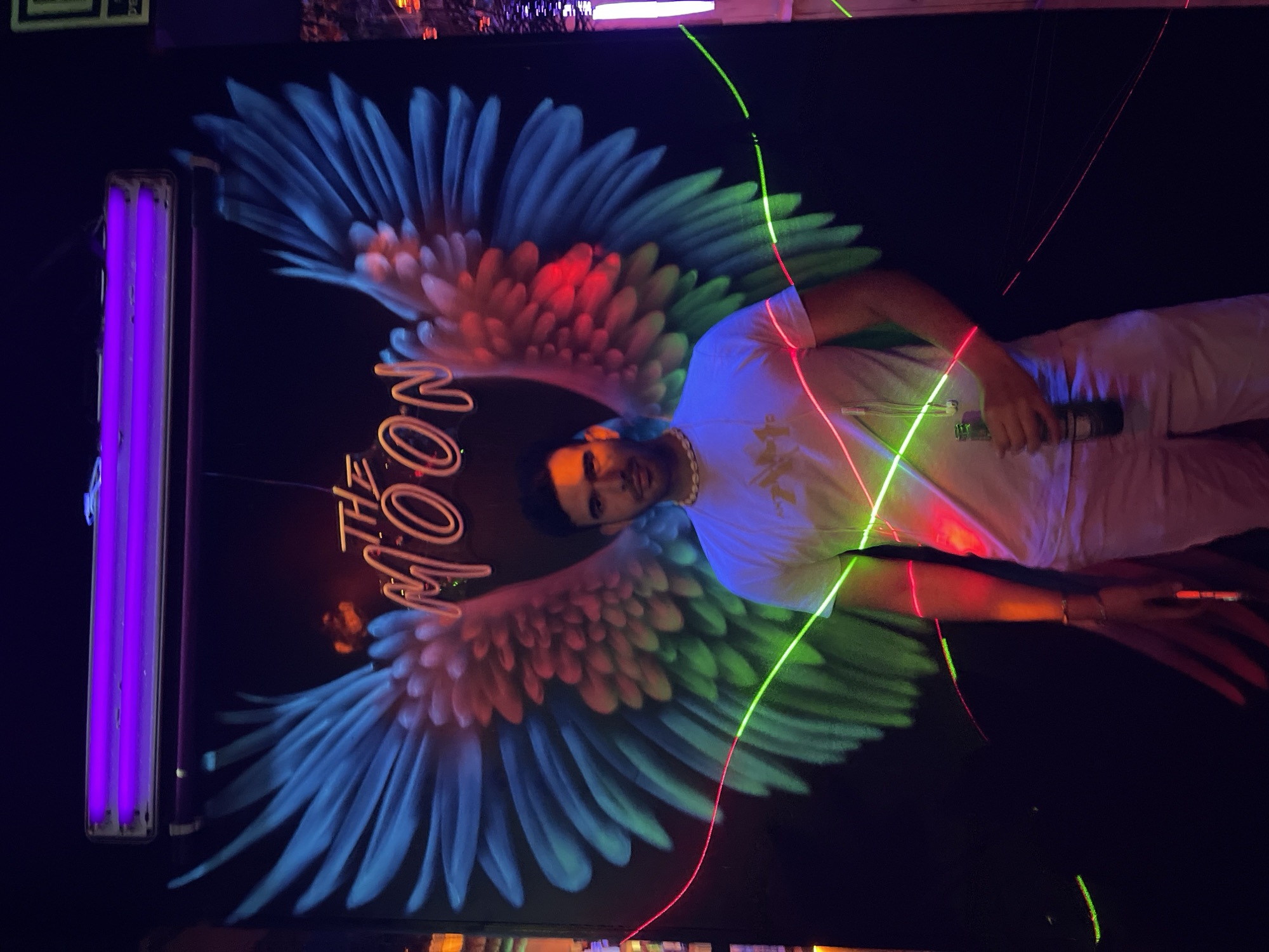A man stands in front of a large mural depicting blue and pink wings with the neon sign "THE MOON" above them. He is wearing a white t-shirt and light-colored pants, with a beaded necklace around his neck and a bracelet on his left wrist. He holds a bottle of beer in his right hand. Neon green and red lights are draped across the scene, creating a vibrant and energetic atmosphere. The background is dark and appears to be a wall. The scene is likely in a nightclub or bar in Barcelona, Spain, given the neon aesthetic and the presence of a bar.