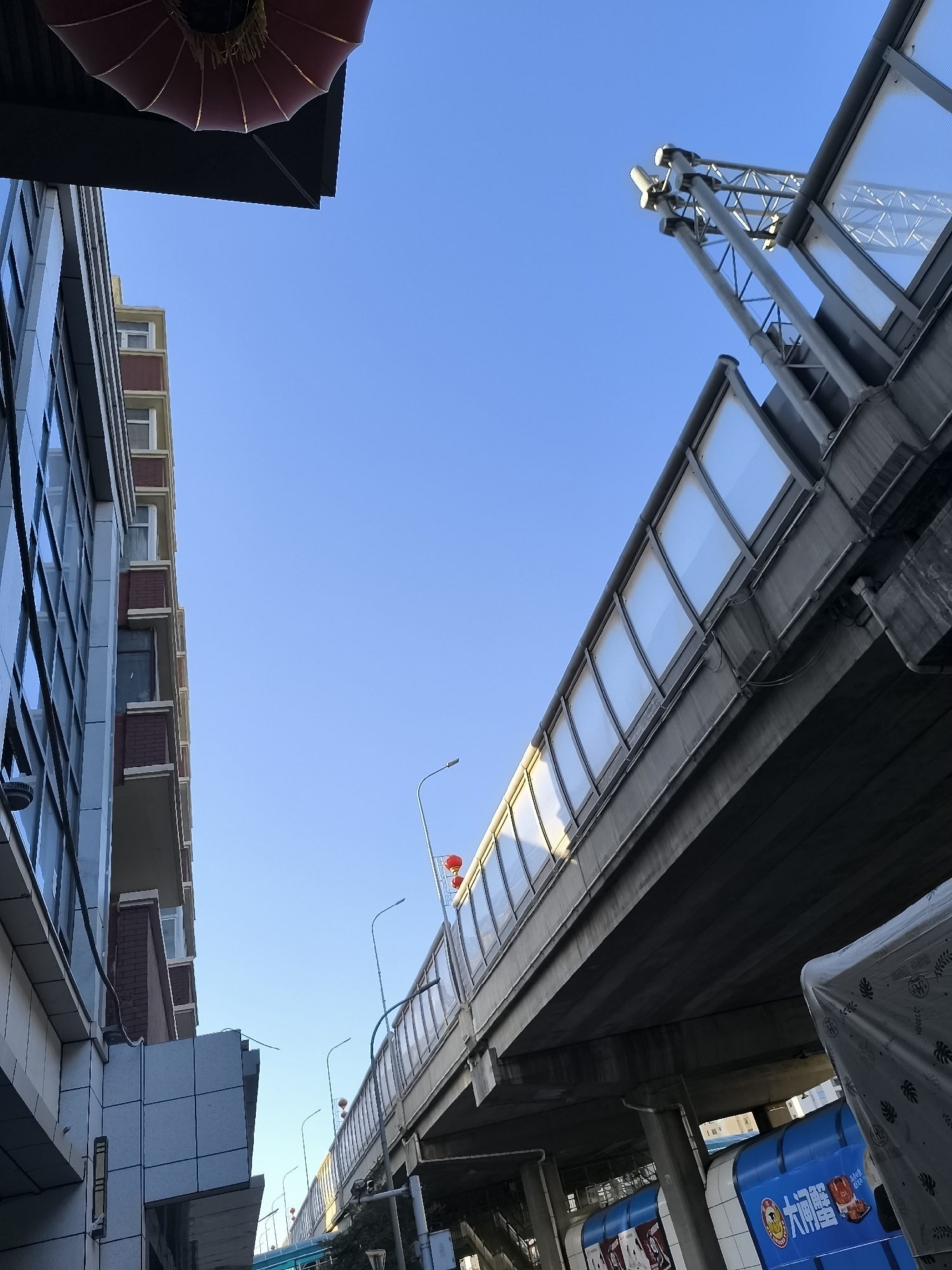 An upward shot captures a clear blue sky above a street-level view of urban infrastructure in Ürümqi, China. To the left, a modern building with a glass facade and brick accents rises vertically. A red Chinese lantern hangs from the overhang of a dark structure at the top left. To the right, a multi-level elevated walkway or bridge dominates the scene, constructed with concrete and featuring a translucent paneling system along its sides. Metal support structures and lighting fixtures are visible on the bridge. Below the bridge, a blue train or subway car with Chinese characters, likely advertisements, is partially visible. The scene depicts a typical urban environment with clear weather conditions.