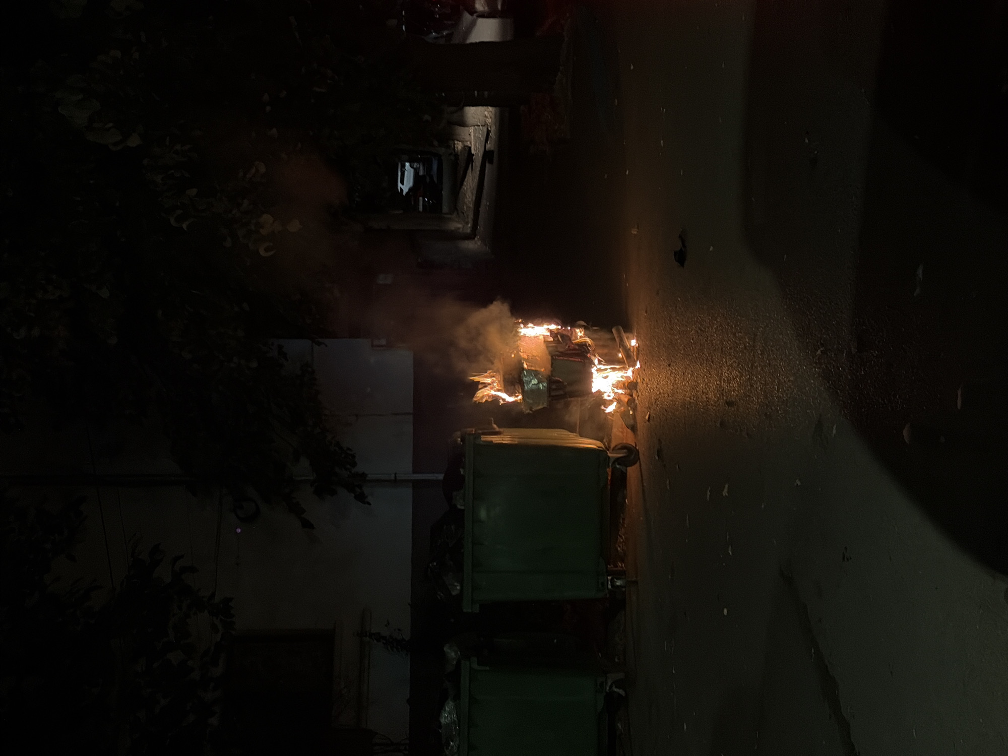 The image depicts an outdoor scene at night, likely in an urban environment. In the foreground, two large, green dumpster bins are visible on the left side of the frame. To the right of the dumpsters, a smaller, rectangular object, possibly a cardboard box, is engulfed in flames and emitting smoke. The fire is bright and casts an orange glow on the asphalt surface. Scattered debris is visible on the ground around the burning object. In the background, trees with dark foliage frame the scene, and a building wall is faintly illuminated on the left. Further in the background, a dimly lit street or alleyway is visible, with a white SUV parked in the distance. The overall atmosphere is dark and somewhat ominous due to the fire and the nighttime setting. The image is set in Vratsa, Bulgaria, suggesting this is a street or alley in that city.