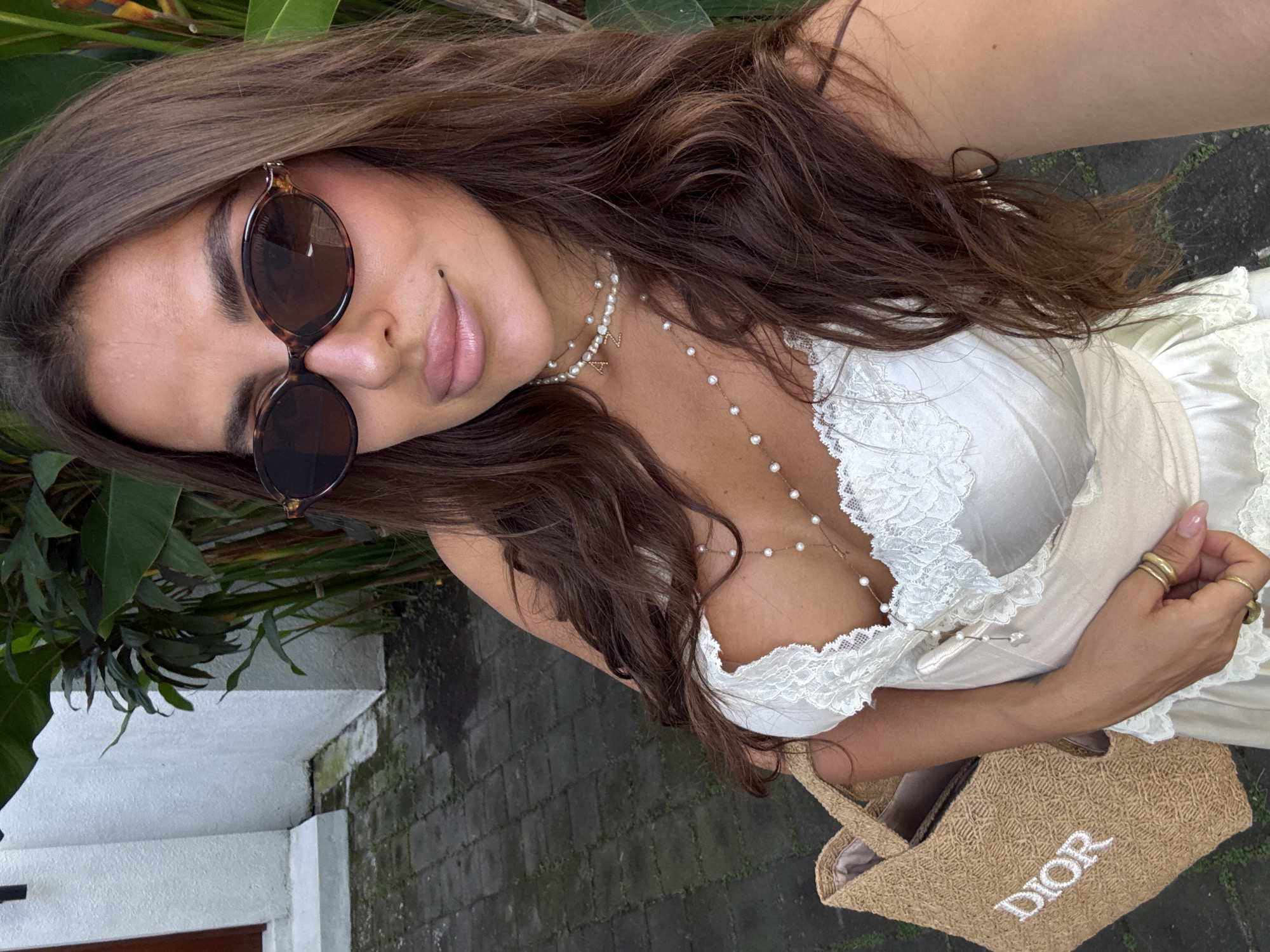 A woman with brown hair and sunglasses is posing in a selfie. She is wearing a white dress with lace trim and a Dior straw tote bag is visible. She is wearing a pearl necklace and rings on her fingers. The background includes foliage, a white building, and a brick walkway. Based on the information provided and the style of the image, this photo was likely taken in North Kuta, Indonesia.