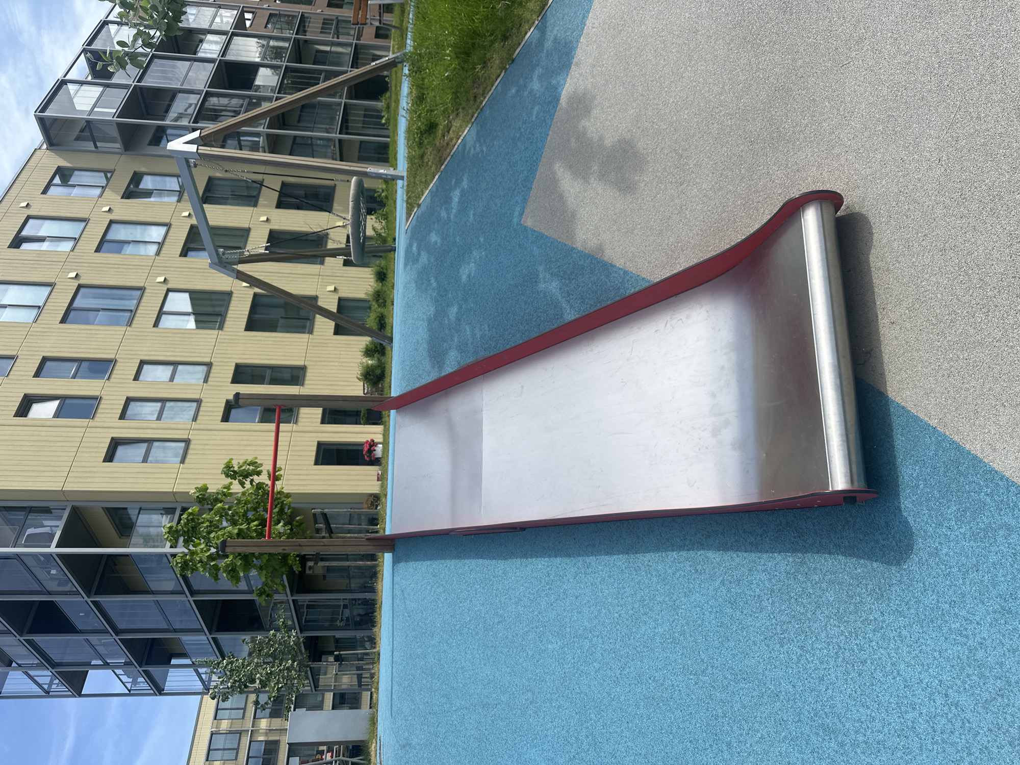 The image depicts a playground slide in front of a modern, multi-story apartment building. The slide is made of metal with red edges and sits on a blue rubberized surface. To the right of the slide, a swing set is visible. The building behind the playground features a light yellow facade with numerous windows and balconies. The scene is set outdoors, under a clear blue sky. This is located in Tallinn, Estonia.