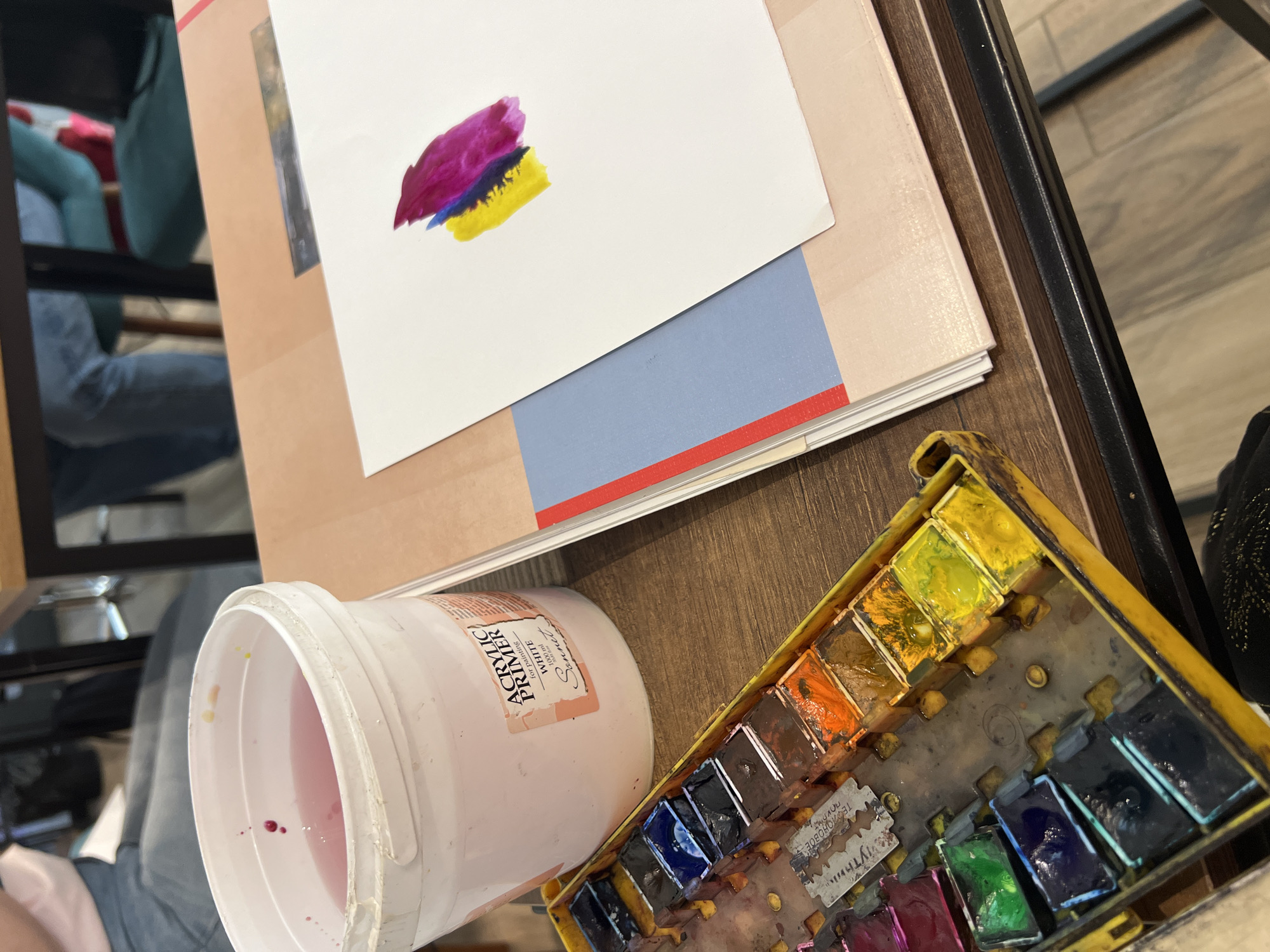 The image presents an overhead view of an art workstation, likely indoors, featuring various art supplies on a wooden table. The main subject is a white sheet of paper with three vivid watercolor swatches—magenta, blue, and yellow—blended at their edges, suggesting an artist is testing colors or beginning a piece. This paper rests on a stack of other boards or papers, one of which has a distinct light blue section and a red stripe.
To the right, a yellow, well-used watercolor palette is open, filled with numerous pans of solid and wet paints in a range of hues including yellows, oranges, blues, greens, and purples. Interestingly, small metallic inserts, resembling repurposed razor blades, are visible within the palette, featuring Cyrillic text such as "ТЕХНОЛОГИЯ" (Technology) and "УЛЫТНЫЙ" (Cozy/Comfortable, possibly part of a brand name).
On the left, a white plastic bucket holds pink-tinted water, indicating its use for rinsing brushes during painting. Beside the bucket is a large white container labeled "ACRYLIC PRIMER" in white, for painting, 500 ml, and the brand "Sonnet," along with additional text in Cyrillic.
In the blurred background on the left, a reflection in a dark, possibly glass or polished surface, reveals a person wearing blue jeans and a blue top, seated and presumably engaged in the art activity. The bright lighting suggests it is daytime. The scene depicts an active artistic process in a creative environment, potentially in Tbilisi, Georgia, given the Cyrillic text on the supplies.