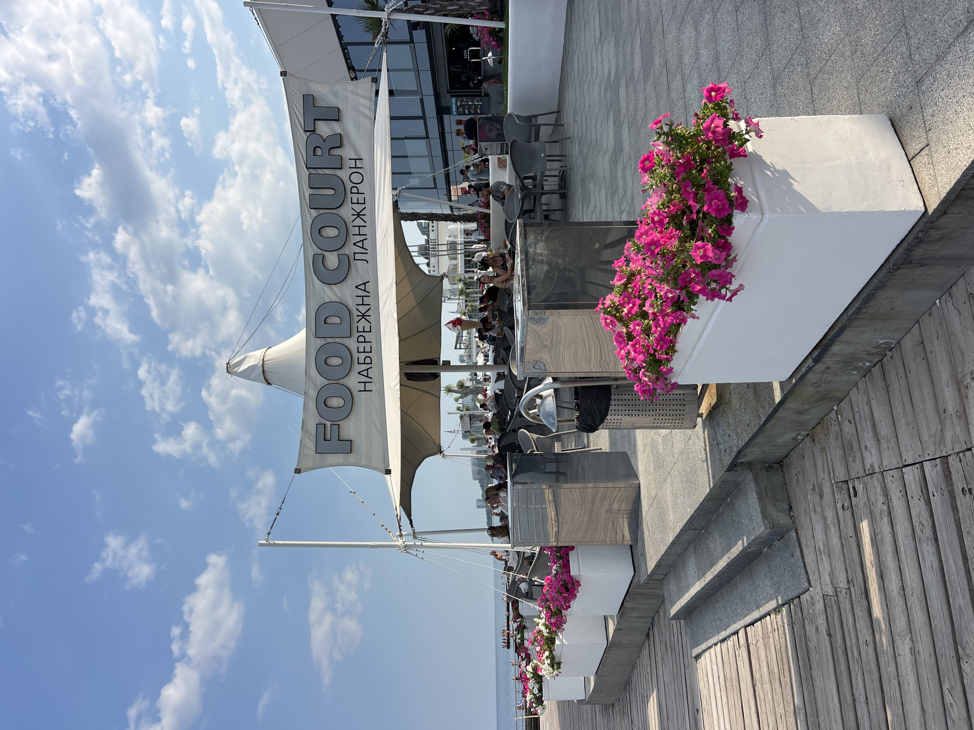 The image depicts a sunny outdoor dining area of a food court situated on the waterfront in Odesa, Ukraine, specifically at Langeron beach. The food court, identified by a banner reading "FOOD COURT" in large gray letters, is covered by a white tent structure supported by poles. In the foreground, a wooden boardwalk leads to a raised area with gray stone steps. Several white rectangular planters filled with vibrant pink petunias are strategically placed along the raised area, adding color to the scene.
Beyond the planters, the dining area is set up with numerous tables and chairs, many of which are occupied by people eating and socializing. Some individuals are seen walking in the background, and the sea is visible to the left, with a clear blue sky and scattered clouds overhead. The architecture of the food court building is modern, featuring large windows. The overall atmosphere is one of a lively, pleasant summer day at a seaside resort.