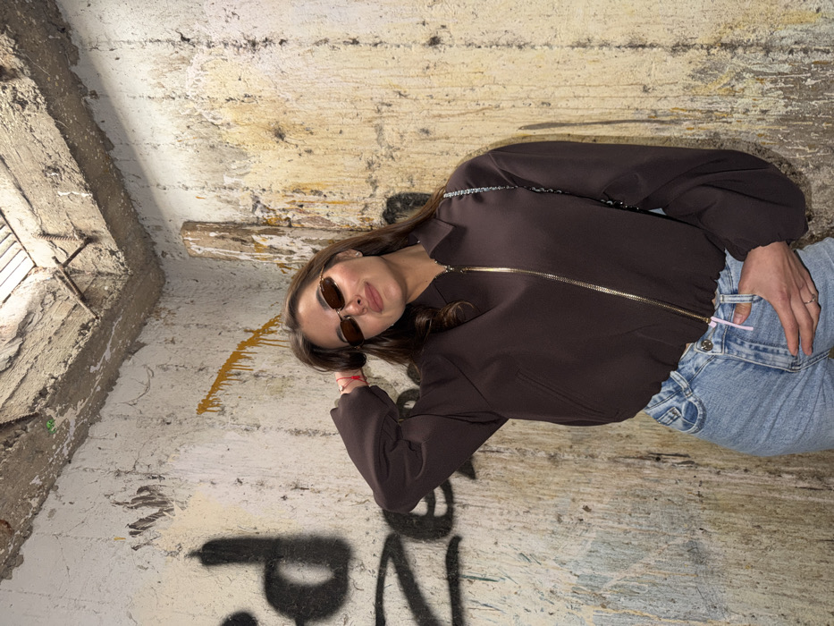 A female subject, appearing to be an adult, is positioned centrally within a corner of a concrete structure. She has long, dark brown hair and wears dark, oval-framed sunglasses, a dark brown zippered bomber jacket with a metallic gold-colored zipper, and light blue denim jeans. A red string bracelet is present on her left wrist, and a thin, metallic chain is visible over her right shoulder. Her left hand is raised, with fingers touching her hair near her left temple, while her right hand rests in the front pocket of her jeans, where a light pink tab or drawstring is visible. She maintains direct eye contact with the viewer, exhibiting a subtle smile.
The environment consists of two intersecting concrete walls and a concrete ceiling, forming a corner. The walls display extensive discoloration, peeling, and staining in shades of yellow, white, and grey, consistent with age and dampness. Black graffiti, partially legible as "ed," is present on the wall to the left of the subject. Vertical streaks of yellow-orange substance are visible on the right wall. The concrete ceiling reveals exposed rebar and a rectangular opening. A raw wooden beam is propped against the corner in the upper right. The scene is brightly lit, likely by an artificial light source such as a camera flash.
This scene is located within the city of Tel-Aviv, Israel, in an environment characteristic of an abandoned, derelict, or utilitarian urban space.