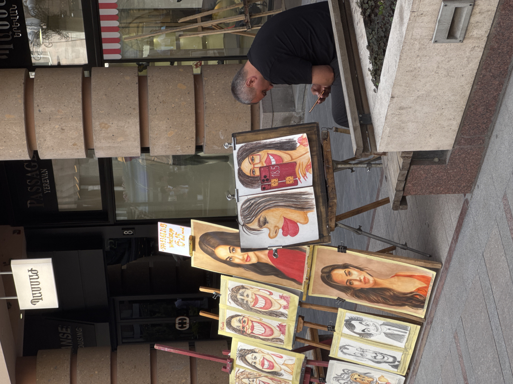 The image depicts a street scene in Yerevan, Armenia. A middle-aged man with gray hair is standing next to an easel, looking at a smartphone in his hand. The easel displays several caricature portraits, including one of a woman holding a smartphone. Other paintings include depictions of women with exaggerated facial features. Next to the easel, there is a sign that reads "37493682493 WhatsApp 15-15 10 minutes DEFAN", suggesting that this is a caricature artist at work. The environment includes a stone planter, a building with the text "PASSAGE YEREVAN" and "ANSE OUSING" visible, and storefront signs in Armenian.