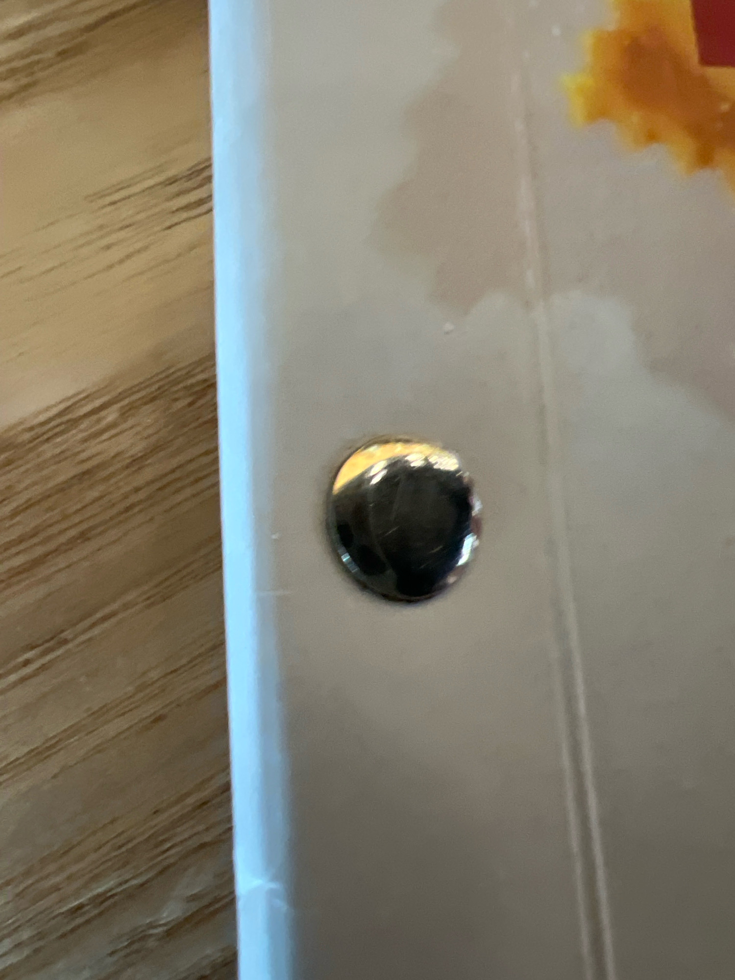 The image is a close-up, eye-level shot of a glossy, white surface with a metallic, circular object embedded in it. The surface appears to be part of a product or packaging, as suggested by a blurred yellow and red shape in the upper right corner. A wooden surface with visible grain is in the background on the left. There are no people or discernible actions taking place. The lighting suggests an indoor setting. There is no information to suggest a location in Krasnoyarsk, Russia.