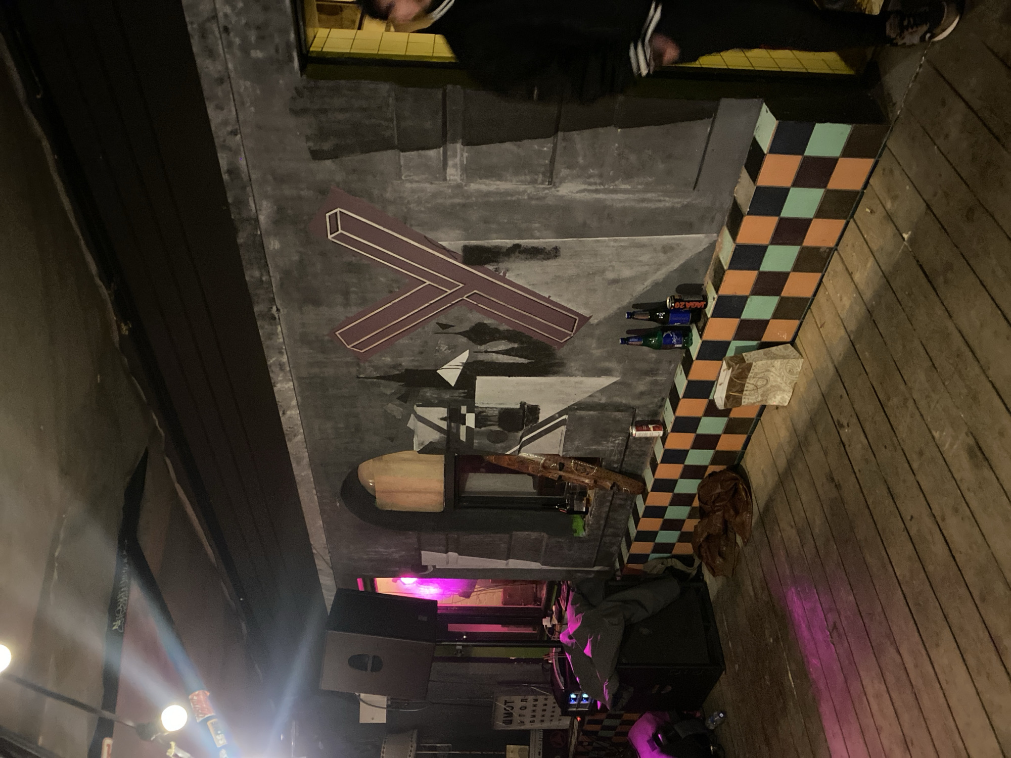 The image shows an interior space, likely a bar or club in Moscow, Russia, given the Cyrillic text on a sign and the general atmosphere. The room has a decorated ceiling with geometric shapes and patterns painted on it. A bar counter with a checkered pattern faces the viewer. Several beer bottles are lined up on the bar top. In the foreground, there is a sound system with speakers and a DJ setup, indicated by the control panel and equipment. Pink and white light emanates from the equipment, illuminating the surrounding area and the wooden floor. A person wearing a black and white Adidas tracksuit is seen in the background near the bar.