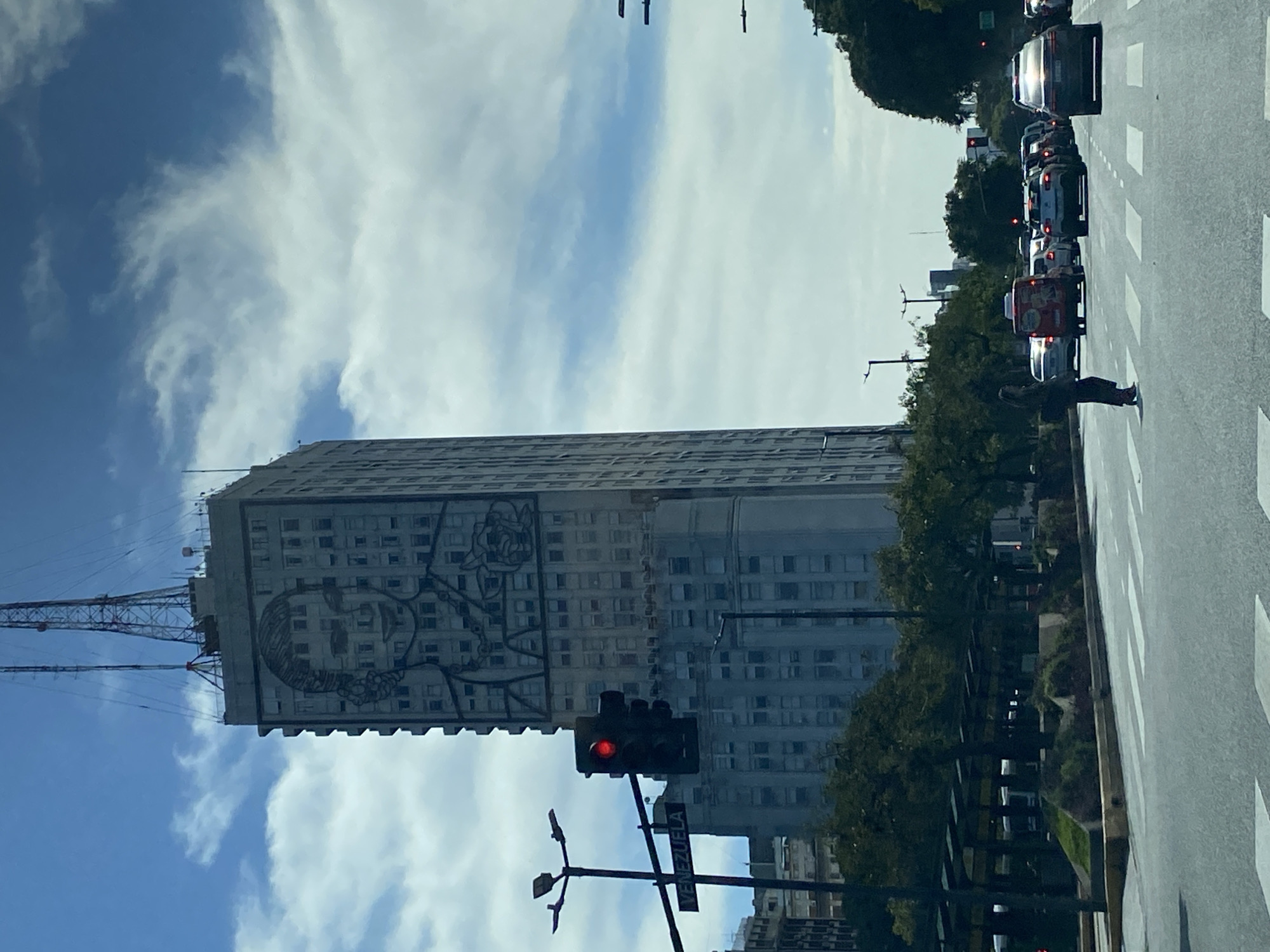 A daytime street scene in an urban environment, likely Buenos Aires, Argentina. A tall, multi-story building dominates the left side of the frame, featuring a large, grayscale mural of a person's face on its exterior. A lattice-style antenna or transmission tower stands on the roof of the building, extending upwards.
To the right of the building, a multi-lane road runs, showing several cars queued, appearing to be stopped at a red traffic light. Headlights and taillights are visible on the vehicles. A pedestrian stands on a white-striped crosswalk on the right side of the road. A red traffic light is prominently displayed in the foreground, with a street sign below it. Trees and other vegetation line parts of the street and are visible in the background, suggesting a green urban space. The sky is blue with scattered white clouds. The street sign clearly displays the text "VENEZUELA."