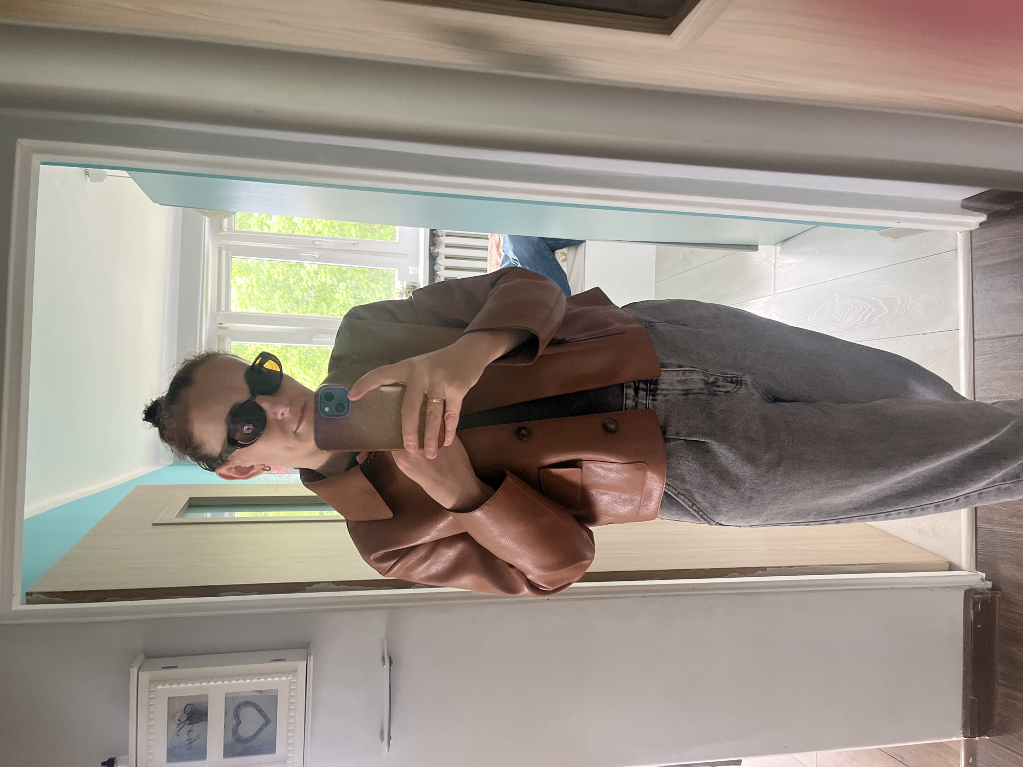 A woman is standing in what appears to be a hallway or entryway, taking a selfie in a mirror. She is wearing sunglasses, a brown leather jacket, and gray jeans. The hallway or entryway has a door leading to a room with a window, allowing natural light to enter. The walls are painted in neutral tones, and there's a decorative picture frame on the wall near the bottom of the frame. The location is in the city of Unknown, Unknown.
