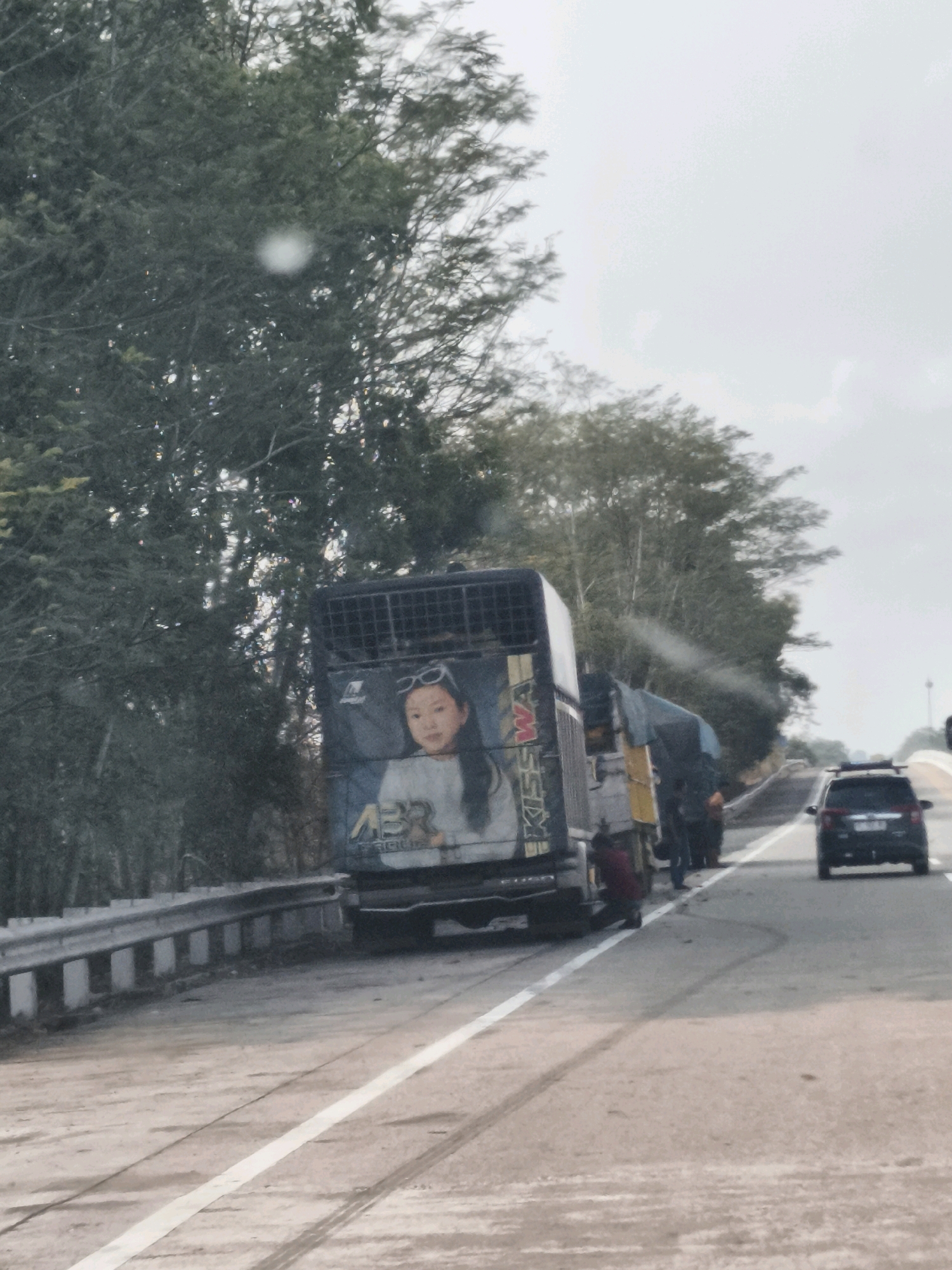 The image shows a side view of a truck on a road, possibly in Lubuk Dalam, Indonesia. The truck has a large mural on its side depicting a young girl wearing sunglasses and a white shirt. The truck appears to be parked on the side of the road, and there are several people standing near it. A black SUV is seen driving on the road in the background. The environment is characterized by trees and vegetation lining the road, with a cloudy sky overhead. The road surface shows tire marks, suggesting recent vehicle activity. A guardrail is visible along the left side of the road.
