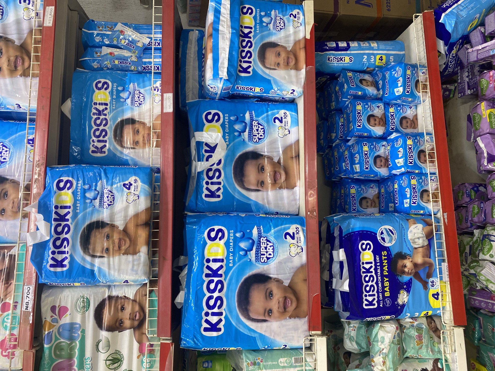 The image depicts a retail display of baby diapers in a store located in Ikorodu, Nigeria. The shelves are stocked with various brands and sizes of diapers, primarily "KissKids" brand. The majority of the products are packaged in blue plastic bags featuring images of babies. Some shelves also show prices, such as "N8,700" and "N11,500". The lighting appears to be artificial, illuminating the products on display. The overall scene is that of a commercial environment focused on baby care products.