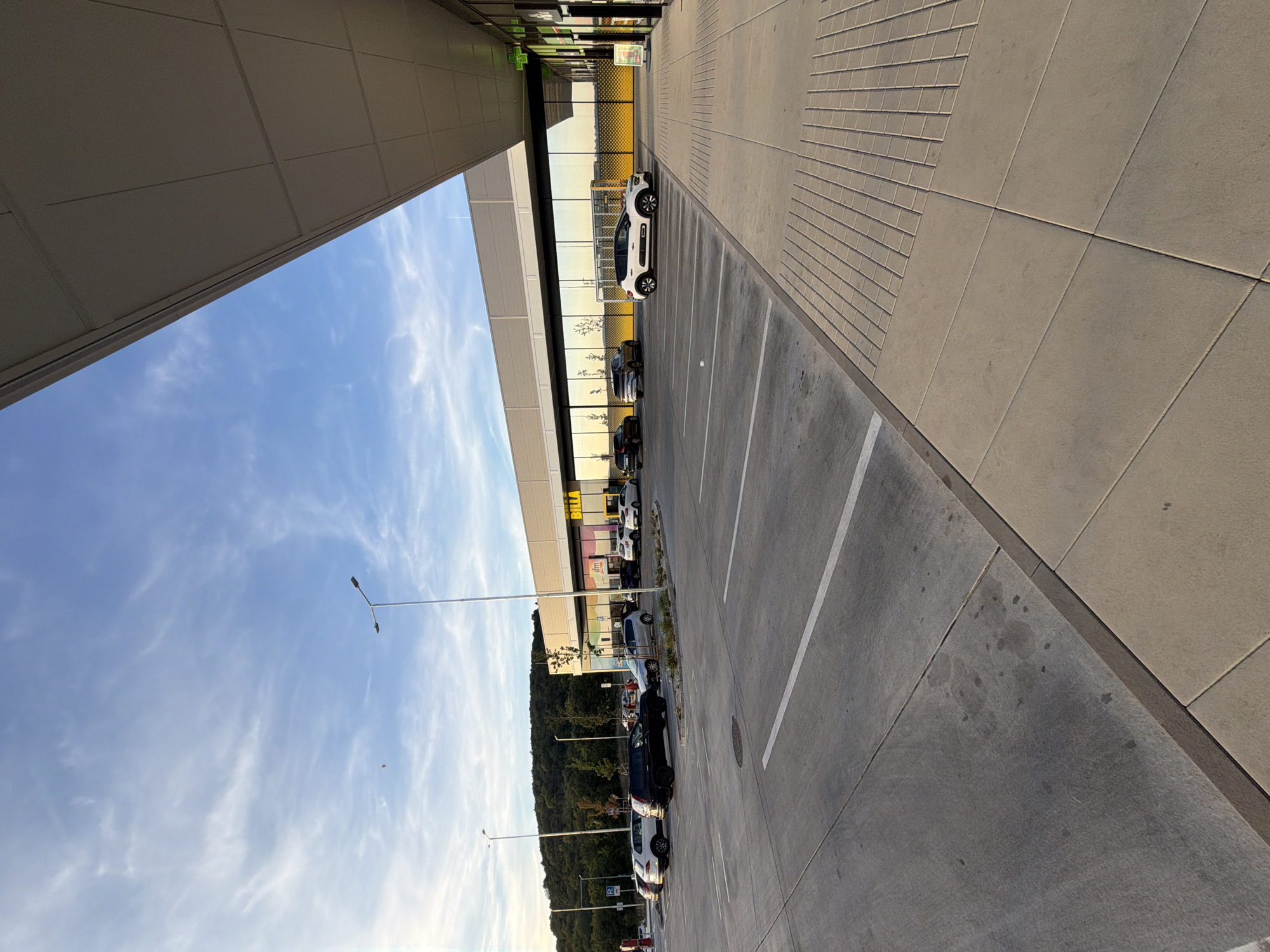 The image depicts a parking lot in front of a commercial building in Nitra, Slovakia. The building features large glass windows and the word "BILLA" is prominently displayed, indicating a supermarket. Several cars are parked in the lot, including SUVs and sedans. The sky is a clear blue with wispy clouds, and the sun is casting long shadows, suggesting it is either early morning or late afternoon. A tree stands near the building, and a hill covered in vegetation is visible in the background. The parking lot is paved with concrete and marked with white lines. A sidewalk made of light-colored pavers runs alongside the parking lot. There are no people visible in the scene.
