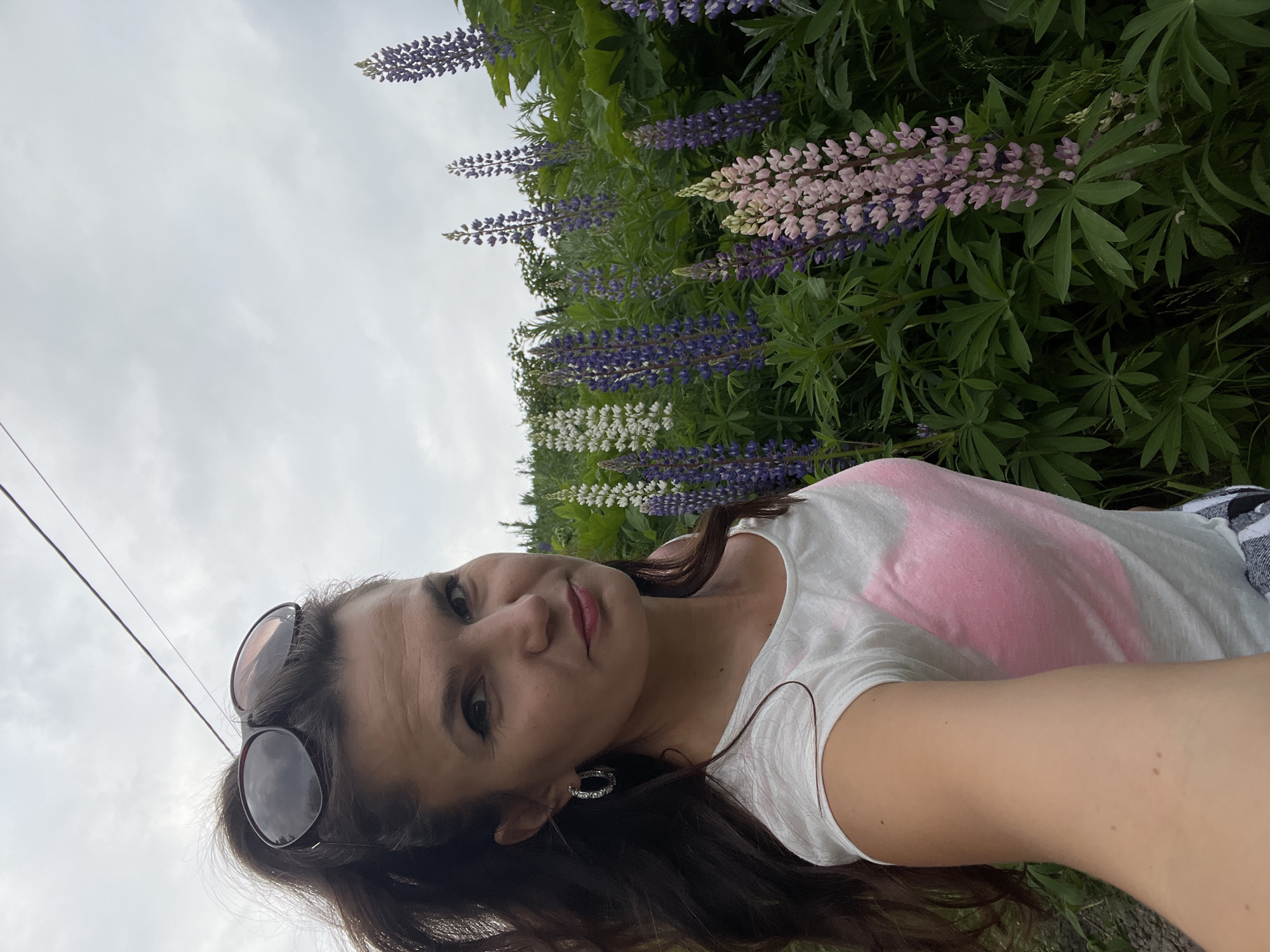 A woman with long, dark hair pulled into a ponytail and wearing sunglasses takes a selfie in a field of lupines. She is wearing a white t-shirt with a pink heart design on the front and has hoop earrings. The lupines are in various shades of purple, white, and pink. The sky is overcast. The scene appears to be outdoors, likely in a rural or park setting. This image does not provide any information that would suggest the location is Petropavlovsk-Kamchatsky, Russia.