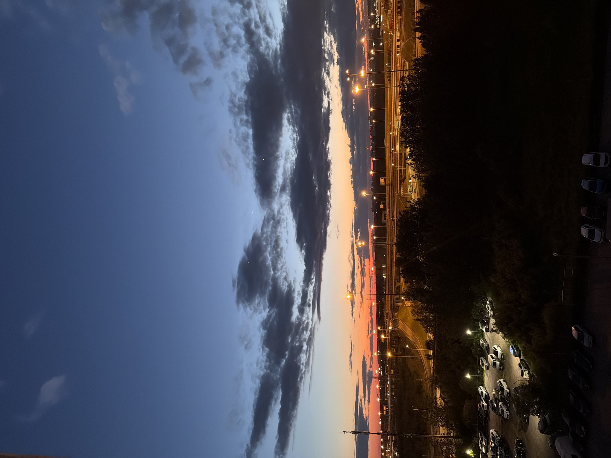 The image depicts a dusk scene in an urban area, likely near a highway or industrial zone. The sky is a gradient of deep blue at the top transitioning to vibrant orange and red near the horizon, indicating the sun has recently set. Dark, scattered clouds are visible against the twilight sky. Numerous streetlights and floodlights illuminate the scene, casting a warm glow on a complex network of overpasses and roadways. A large parking lot, filled with numerous cars, is situated in the foreground and middle ground. Trees and dark foliage border the parking lot and the roadway, creating a distinct separation between the man-made infrastructure and the natural elements. The overall impression is one of an active, illuminated urban landscape transitioning into night.