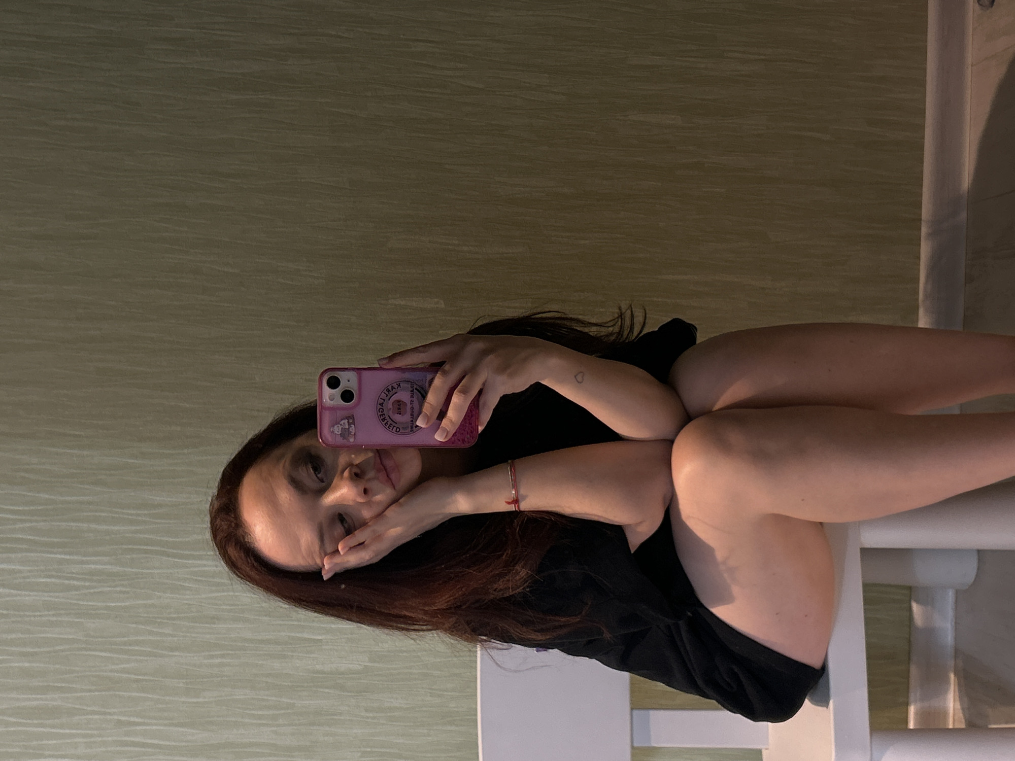 A woman is taking a selfie while sitting on a white chair or structure. She has long, dark brown hair and is wearing a black top. She is holding a pink phone case. The background appears to be a light beige wall. It is not possible to definitively confirm the location as Chaikovsky, Russia from the given image.
