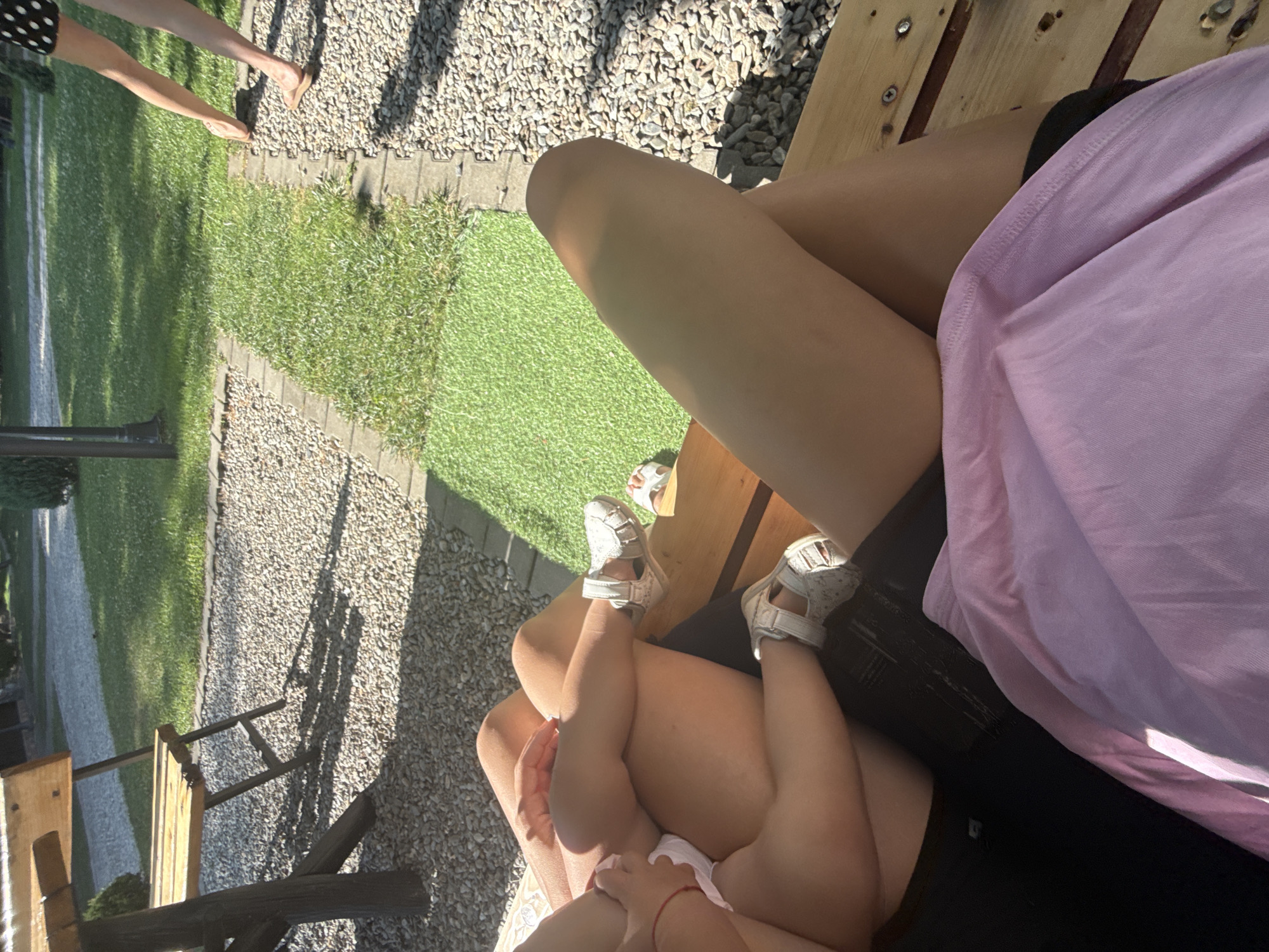 The image captures a close-up view of legs and feet, set against an outdoor environment with a gravel path and a patch of green grass. In the foreground, a person's legs are crossed, wearing black shorts and a pink top. To the left, a child's legs are visible, adorned with white sandals, resting on another person's lap or a similar surface. The child is also wearing a white garment. Further in the background, another individual's legs are seen, one of which is wearing a sandal, walking on the gravel. The scene appears to be a casual outdoor setting, possibly a backyard or a park area, with a wooden bench or table structure in the foreground. There is a wooden bench visible in the background. The setting is likely Cârțișoara, Romania, given the provided context.