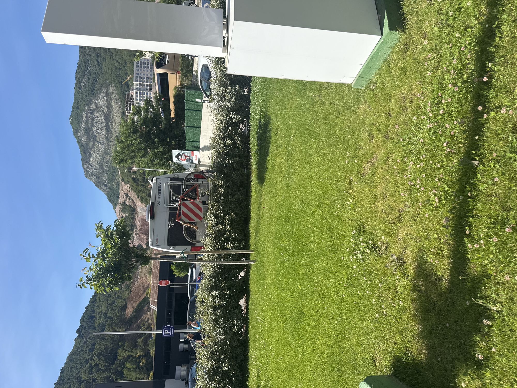 The image depicts a grassy area with a partial view of a city street and mountains in the background. The location is Budva, Montenegro.
In the foreground, there's a patch of green grass with small pink and white flowers. A trimmed hedge divides the grass from the street. Behind the hedge, a white camper van with a bicycle rack is parked. The van has a red and white striped hazard sign.
Beyond the van, there is a parking area with cars and parking signs. In the background, there's a mountain and some buildings, indicating the surrounding landscape of Budva. The sky is clear and blue.