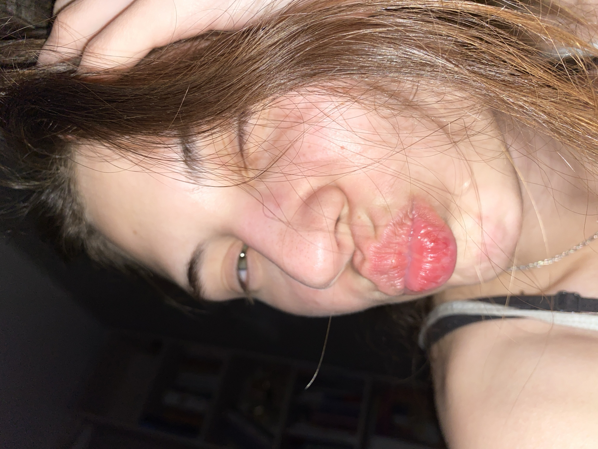 A close-up, rotated image features a person with fair skin and light brown hair. Their hair is partially obscuring their face, with their fingers running through it. The person's lips are puckered. A single strand of saliva hangs from their bottom lip. They are looking downward and to the left. A silver necklace is visible on their neck. The person wears a black and white striped tank top. The background is dark and partially out of focus, revealing a shelf. The location of this image is unknown and is not verified to be in Votkinsk, Russia.