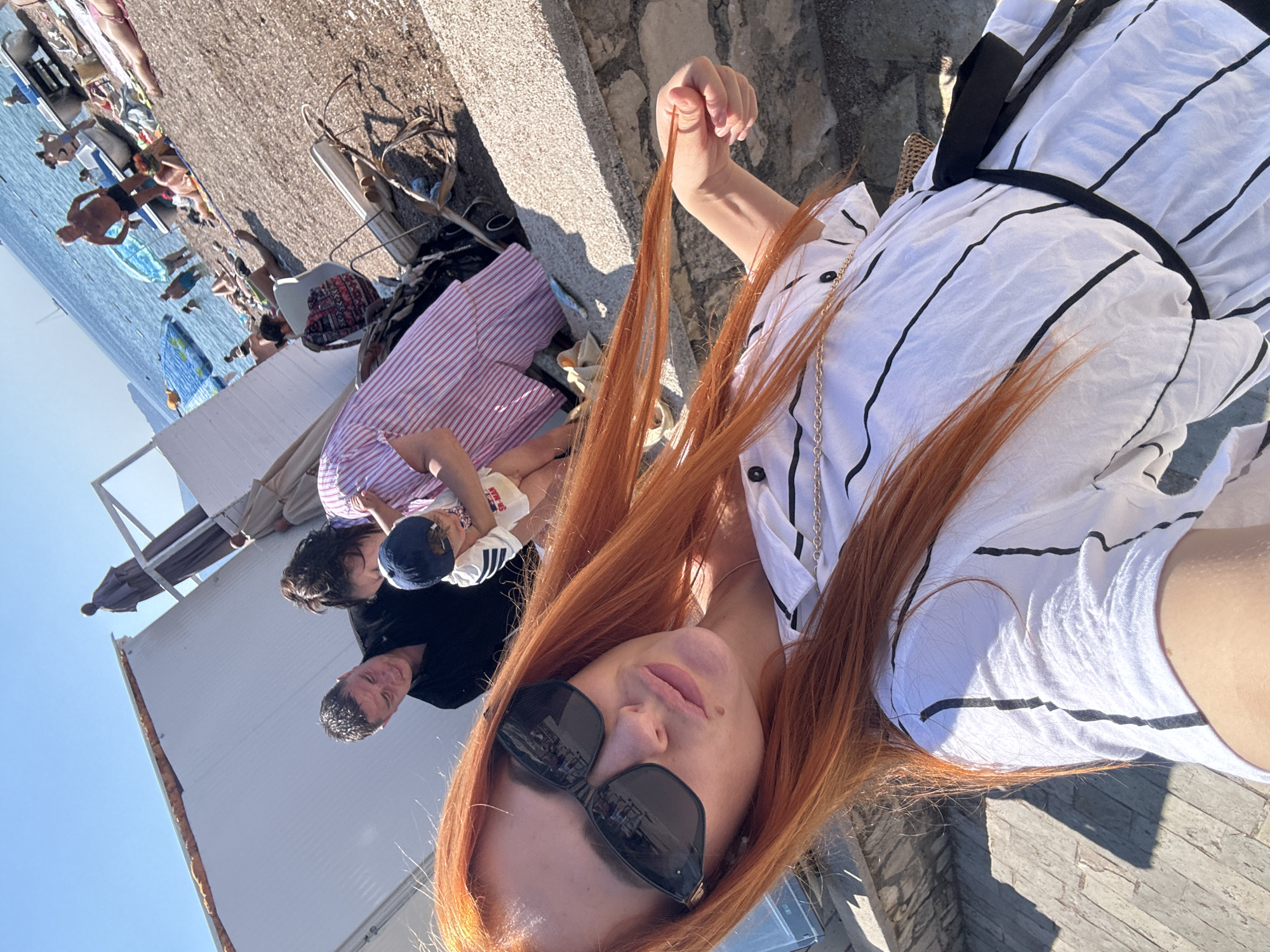 This is a vertical, eye-level, selfie-style photo taken on a sunny day, likely late morning or early afternoon, judging by the bright sunlight and sharp shadows. The scene is a crowded beach in Budva, Montenegro, with a clear blue sky overhead.
The primary subject is a woman with long, auburn hair, wearing a white and black striped shirt and large dark sunglasses. She is holding a strand of her hair, and her face is turned towards the camera.
In the background, a diverse group of people are visible, suggesting a lively beach atmosphere. Some are lying on lounge chairs or towels on the pebble beach, while others are closer to the water, which is a striking blue. There are beach umbrellas and bags scattered around.
Several other people are in the foreground and mid-ground of the image. To the left of the woman, partially visible, are two children and an adult man. One child is wearing a blue cap and a white shirt with stripes, and the other child appears to be lying down. The adult man is wearing a dark t-shirt and has short, dark hair. A woman in a pink and white striped top is also visible.
The setting includes a concrete or stone walkway behind the people on the beach, and further back, what appears to be a white structure, possibly a building or a beach cabana. There is no visible text in the image. The overall mood is relaxed and summery, capturing a moment of leisure at the beach.