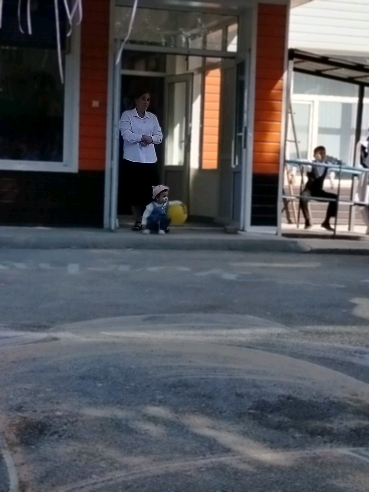 The image depicts an outdoor scene, possibly near a building with orange and white siding and glass doors. A woman in a white collared shirt and dark skirt stands near an entrance. In front of her, a young child wearing a pink hat and blue overalls is sitting on the ground. To the right of the entrance, another child is visible behind a railing, and a ladder is propped against the building. The ground in the foreground appears to be paved with a textured surface. The overall setting suggests a public space or the exterior of a community building in Көлкент, Kazakhstan.