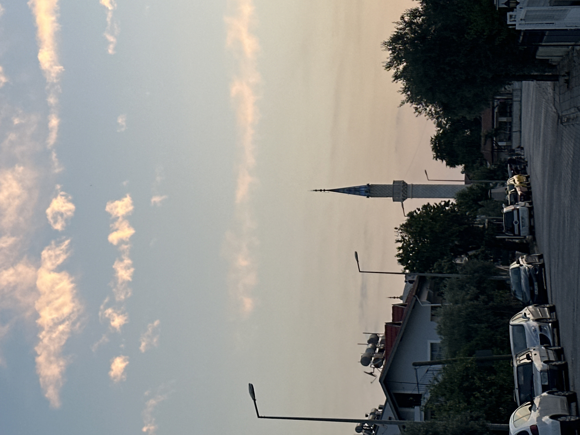 The image depicts a street scene at dusk in Fethiye, Turkey. A mosque with a tall, slender minaret is visible in the background, its top silhouetted against the pale pink and blue sky. Several cars are parked along the side of the paved street. Houses with red-tiled roofs and solar water heaters are also present. Tall streetlights line the road, and lush green trees and bushes frame the scene. The sky is adorned with wispy, pale orange and pink clouds, suggesting the last moments of sunset. The overall impression is one of a quiet, residential area settling in for the evening.