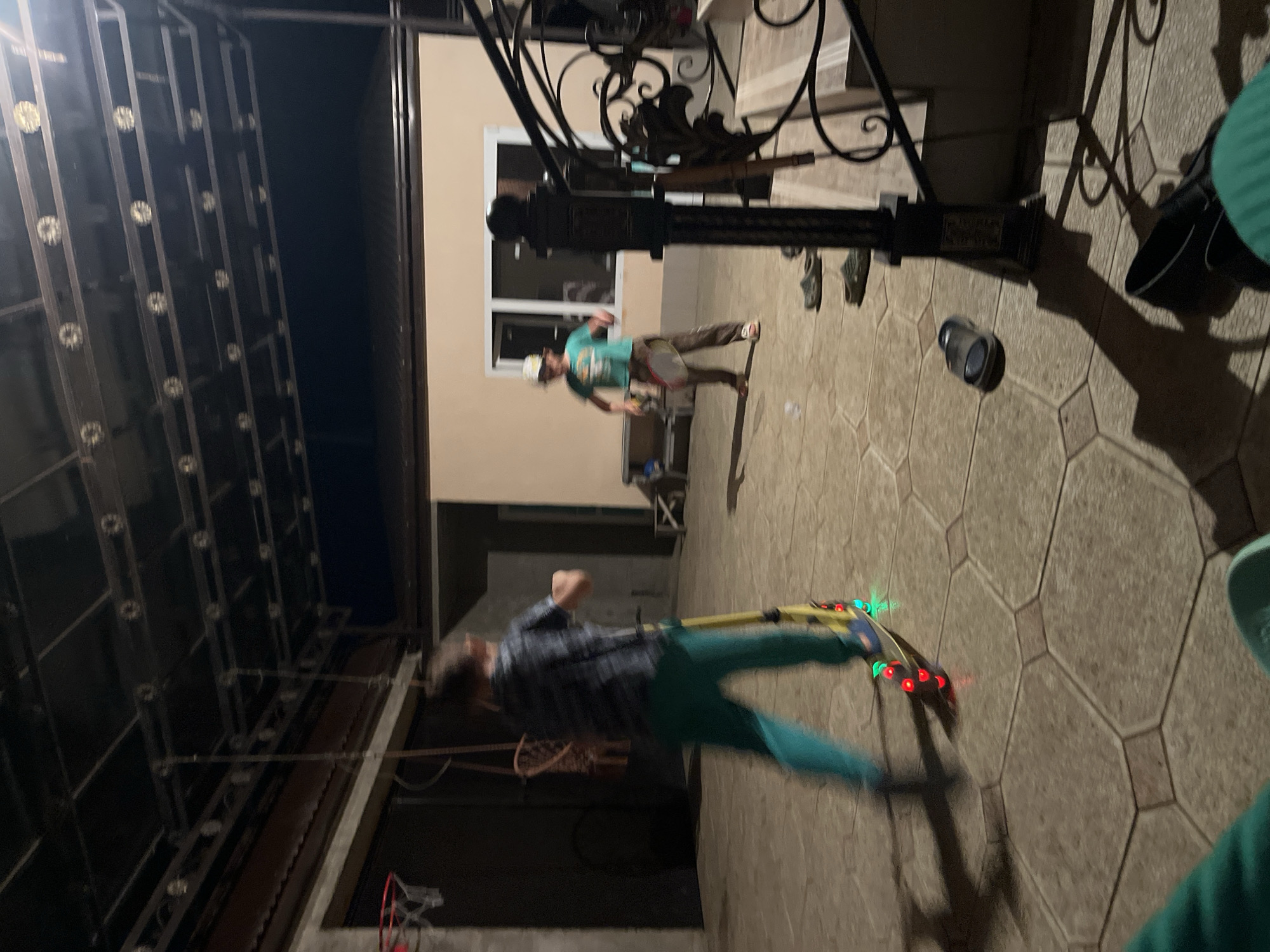 This image depicts a nighttime scene on a paved outdoor area, likely a courtyard or patio, with a building in the background. Two young boys are present. One boy is on the left, blurred due to motion, riding a yellow scooter with illuminated wheels. He is wearing a dark, patterned t-shirt and teal pants. The other boy, on the right, is also blurred and appears to be playing with a racket and possibly a shuttlecock. He is wearing a light green t-shirt, brown pants, and a white baseball cap. The environment includes a dark ceiling structure with lighting fixtures above, a basketball hoop mounted on a wall to the left, and a set of stairs with an ornate black metal railing on the right. Several pairs of sandals are scattered on the ground. The overall impression is a casual, recreational activity taking place outdoors after dark. The location is identified as Kaspiysk, Russia.