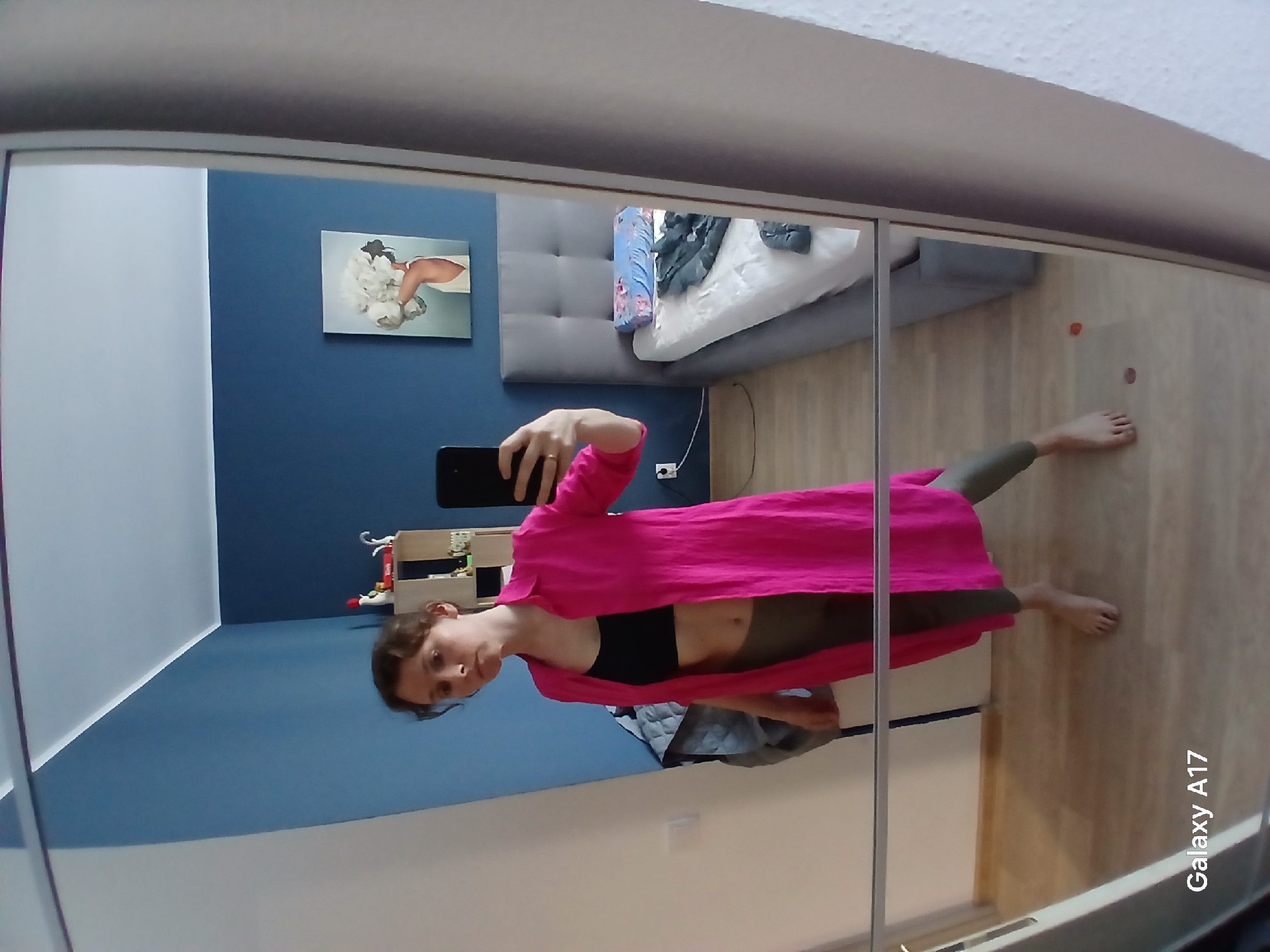 A person is reflected in a mirror. The person is holding a phone and wearing a pink top, black top, and olive green pants. The background includes a blue wall with a picture, a bed with pillows and blankets, a wooden floor, and a toy shelf. The reflection also shows the side of a wardrobe. The camera model "Galaxy A17" is printed on the lower right corner of the image. The location of the scene in the city of Yekaterinburg, Russia is not evident from the image.