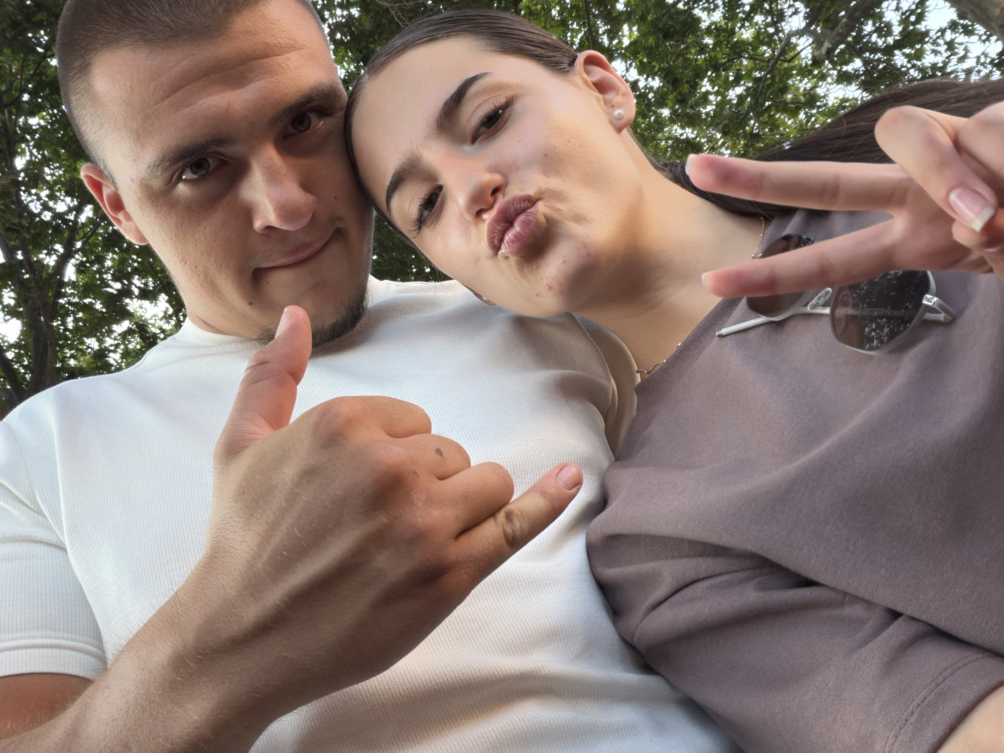 Two individuals, a male and a female, are captured in a close-up, low-angle selfie. The male, with dark hair and a beard, is positioned behind the female, looking towards the camera. He is wearing a white t-shirt. His left hand is extended towards the camera, with his thumb and index finger pointing downwards.
The female, with dark hair pulled back, is in the foreground, facing the camera with her lips puckered. She is wearing a mauve t-shirt and has a pearl earring in her right ear. She is holding up two fingers in a peace sign with her right hand. A pair of sunglasses is hanging from her t-shirt.
The background consists of green foliage, suggesting an outdoor setting, possibly in a park or garden. The lighting indicates it is daytime. No specific location in Burgas, Bulgaria, can be determined from the image.