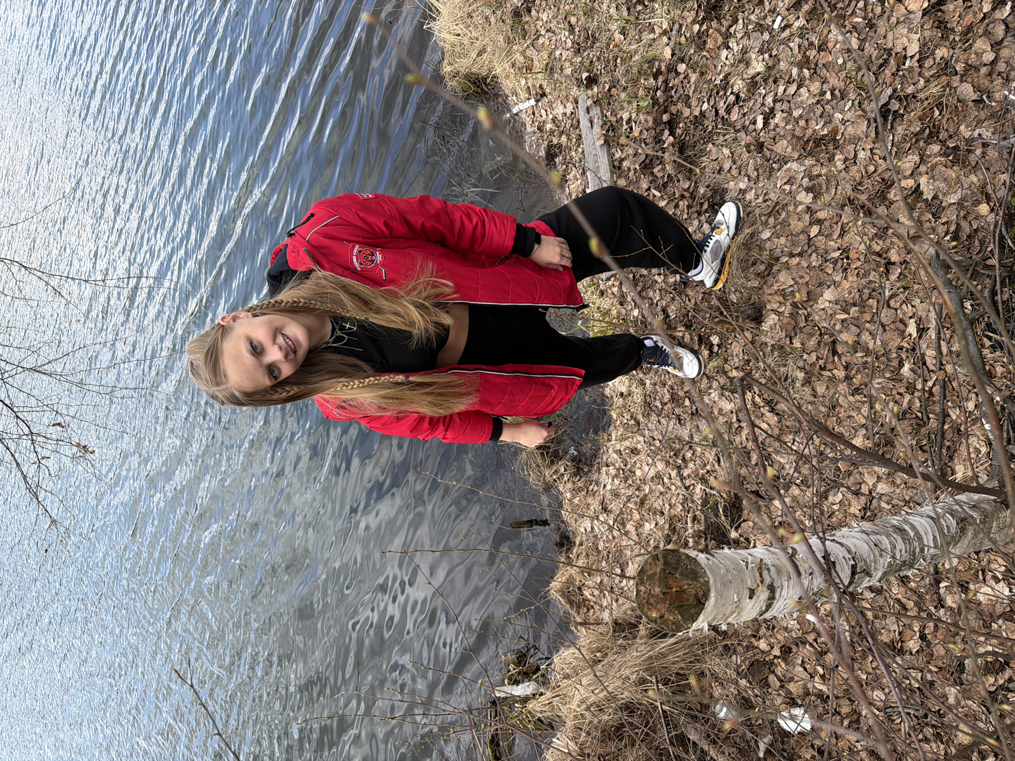 A young woman with long blonde hair styled in braids stands next to a body of water. She is wearing a red jacket, black top, and black pants, along with white and black sneakers. She is standing on a shore covered in fallen leaves and sparse vegetation. The water is reflecting the sky. A cut birch log lies on the ground near her. The scene appears to be in a natural area near a lake or river. The location is Novyi, Russia.
